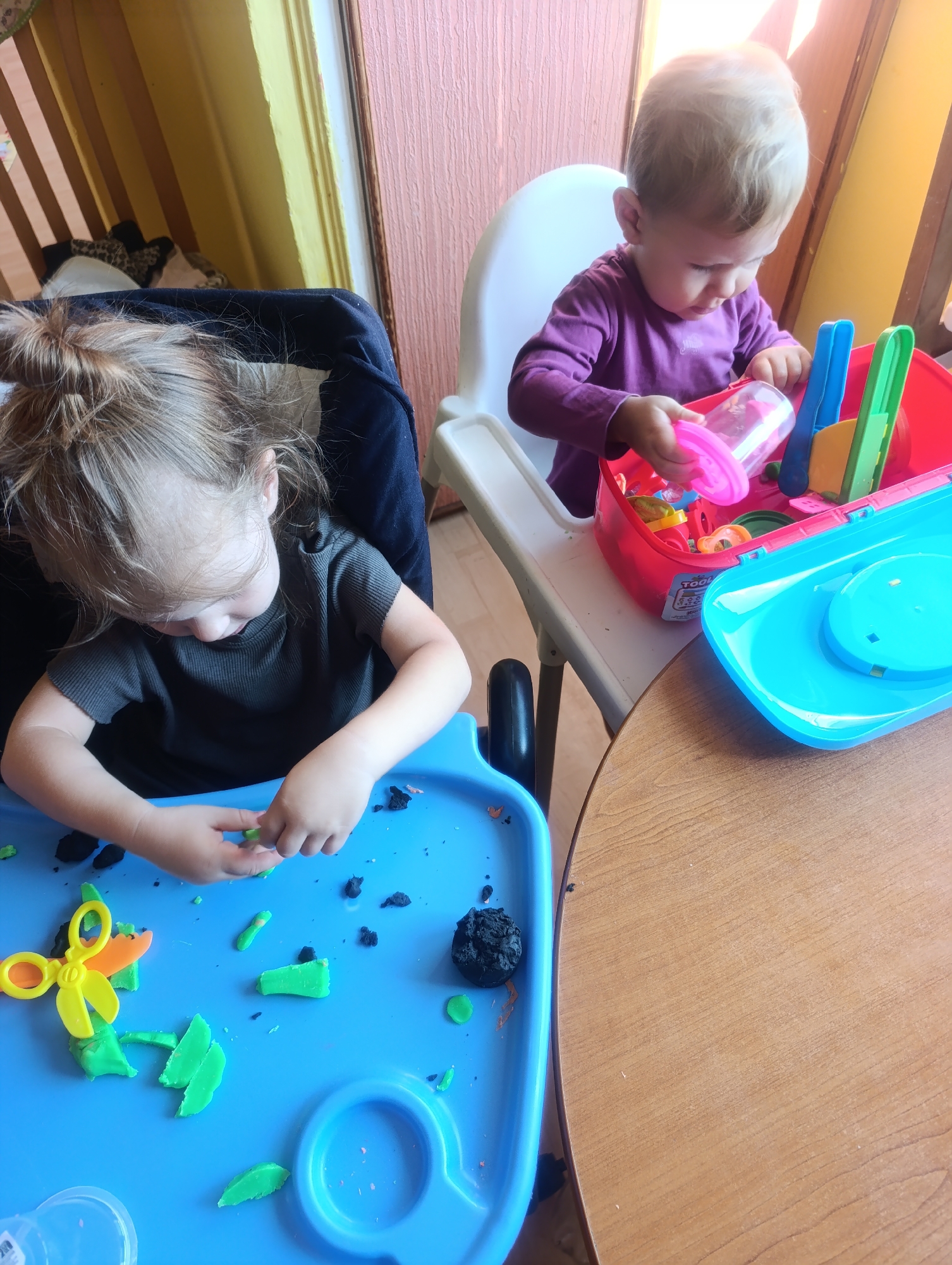 In a domestic setting in Olomouc, Czechia, two young children are engaged in playing with playdough. The child on the left, seated in a high chair with a blue tray, is actively manipulating green and black playdough. A pair of yellow plastic scissors is also visible on the tray, alongside various playdough shapes. The child on the right, seated in a white high chair, is reaching into a pink playdough container filled with colorful tools. The background includes a wooden crib with a stuffed animal, and a pink and yellow wall. The overall scene depicts a typical playtime activity for young children.