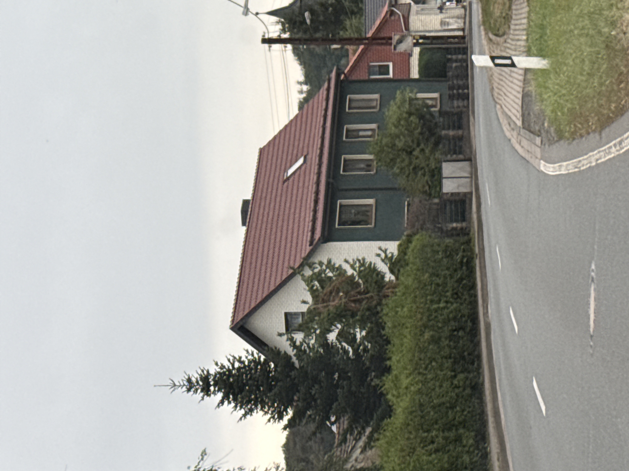The image shows a street scene in the city of Ellrich, Germany. A two-story house with a red tiled roof and white upper half is partially visible on the right side of the frame, bordering a dark green section. Adjacent to this house, another building with a red tiled roof and white siding is visible. A dark green hedge lines the left side of the road, behind which more trees and foliage can be seen. The road itself is asphalt with white lane markings. A white and black directional post stands on the grassy shoulder of the road on the right. The sky is overcast.