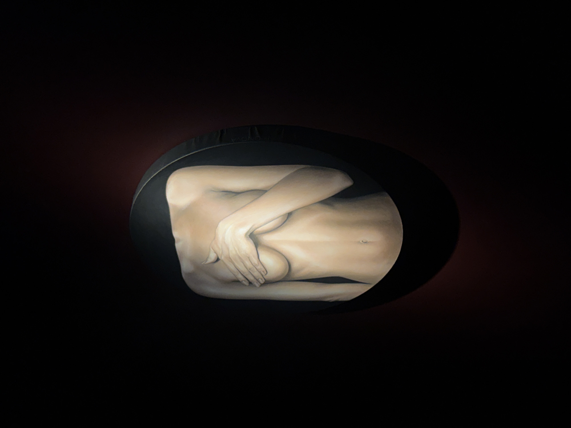 This media file presents a realistic painting or digital artwork of a human torso, likely female, from the chest to the upper thighs. The figure is depicted in a curled or fetal-like posture, with light-toned skin prominently highlighted. Her arms are crossed over her chest, and her hands gently cover her breasts. The body is contained within a dark, elliptical, or egg-shaped vessel, which serves as a central frame. This vessel is set against an almost black background with subtle hints of a dark reddish-brown hue, particularly towards the upper right. The lighting on the figure is soft and emphasizes the smooth contours of the body, creating a strong contrast with the dark surroundings. The overall scene suggests introspection, vulnerability, and containment. Faint white text, possibly a signature, reading "YOKAI" is visible on the upper edge of the dark elliptical vessel. The art piece is located in Moscow, Russia.