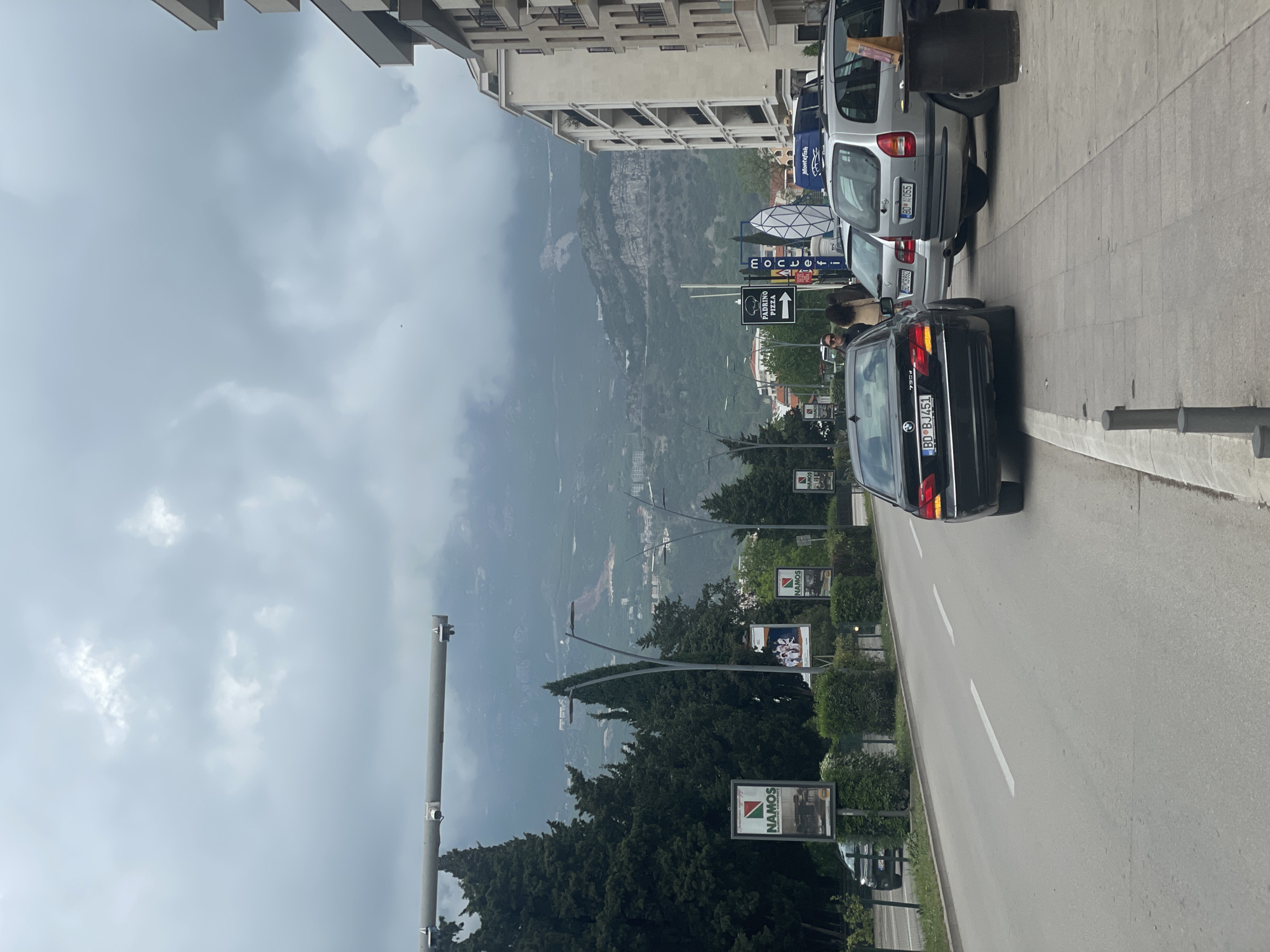 This media file captures a street scene in Budva, Montenegro, on an overcast day. The image is framed with a wide road running through the middle foreground, separating a sidewalk with parked cars on the right from buildings and a natural landscape on the left.
In the background, majestic mountains rise, partially obscured by clouds or haze, indicating their significant elevation. Closer to the viewer, the urban environment consists of multi-story buildings, some with visible balconies, lining the street. Lush green trees and trimmed bushes are prominent along the roadside and in the mid-ground, contributing to the natural elements within the town setting.
On the right side of the road, several cars are parked alongside a sidewalk. A black BMW sedan, model 730d, is visible in the foreground, displaying a license plate "BD BJ451". Behind it, a silver minivan with the license plate "BD 0G65" is parked, followed by another silver car with "BD 0G6Z". Two individuals are partially visible near the parked cars, one with dark, curly hair, suggesting pedestrian activity.
Various commercial signs are scattered throughout the scene. Notably, a "PADRINO PIZZA" sign with a directional arrow is visible, along with signs for "MonteFish" and "MOJ CVJECAR". Multiple billboards advertise "NAMOS Real Estate Company". Streetlights and utility poles are also present.
The sky is heavily overcast with prominent grey clouds, suggesting either an early morning, late afternoon, or generally cloudy day. The overall impression is that of a typical, active street in a town nestled against a mountainous backdrop.