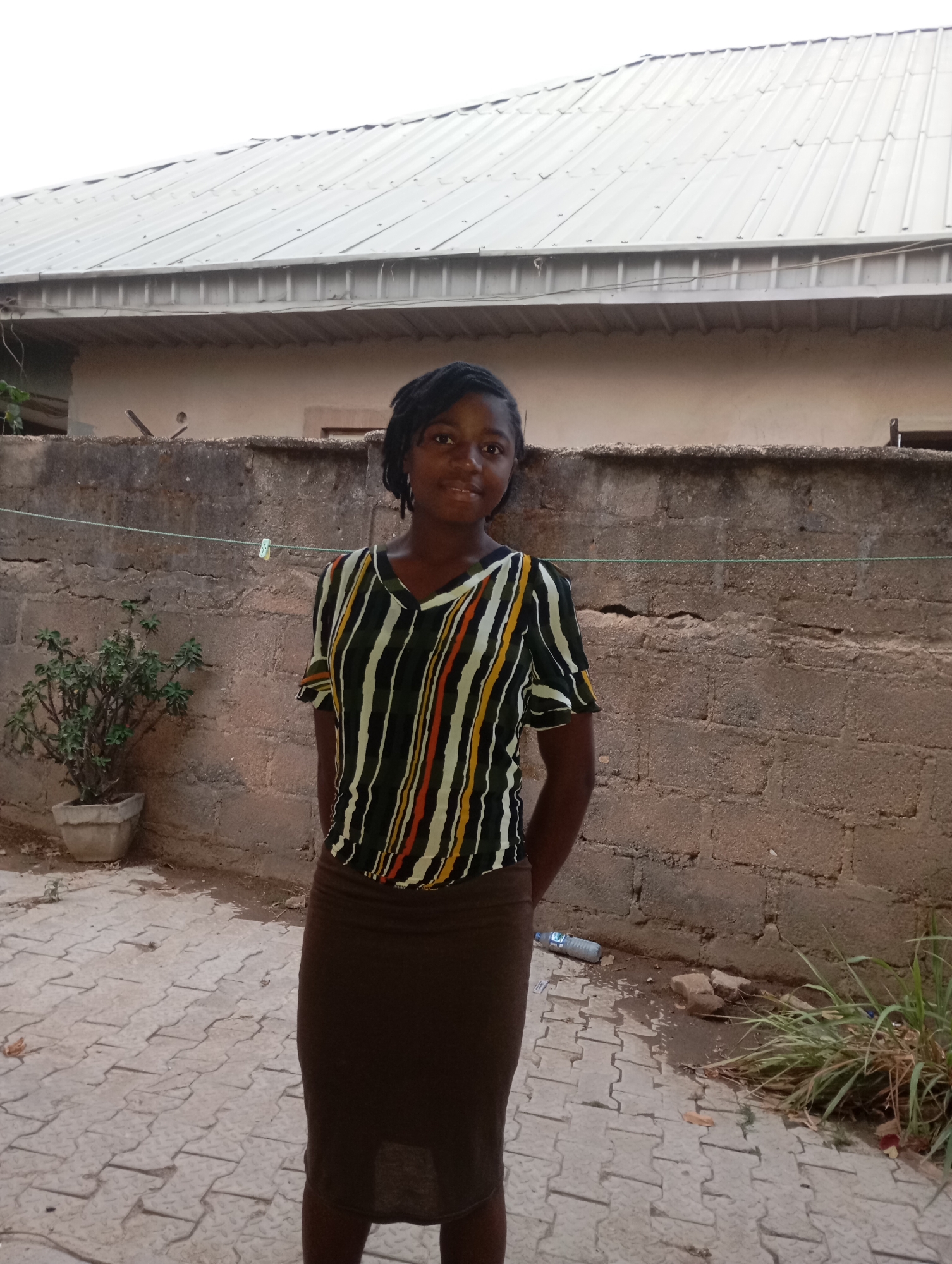 A young female stands centrally in an outdoor paved area, posing for a photograph. She has dark skin and dark hair, possibly styled in braids or dreadlocks, pulled back from her face. She wears a short-sleeved V-neck top with vertical stripes in black, white, orange, yellow, and green, paired with a dark brown, knee-length skirt. Her hands are clasped behind her back, and she exhibits a calm, faint smile.
Behind her, a weathered concrete block wall extends horizontally across the midground. A thin green string, possibly a clothesline, is strung along the wall, with one white clothespin visible. Above the wall, the light grey corrugated metal roof of a building is visible, with a lighter-colored building wall beneath it. To the left of the young woman, a small green plant grows in a light-colored concrete pot. To her right, near the base of the wall, is a patch of green grass and some scattered debris, including a clear plastic water bottle and small rocks. The ground she stands on is paved with grey interlocking concrete blocks arranged in a pattern. The bright, even lighting suggests it is daytime. This scene likely depicts a casual outdoor setting, such as a yard or compound, in Jos, Nigeria. No discernible text is visible in the image.