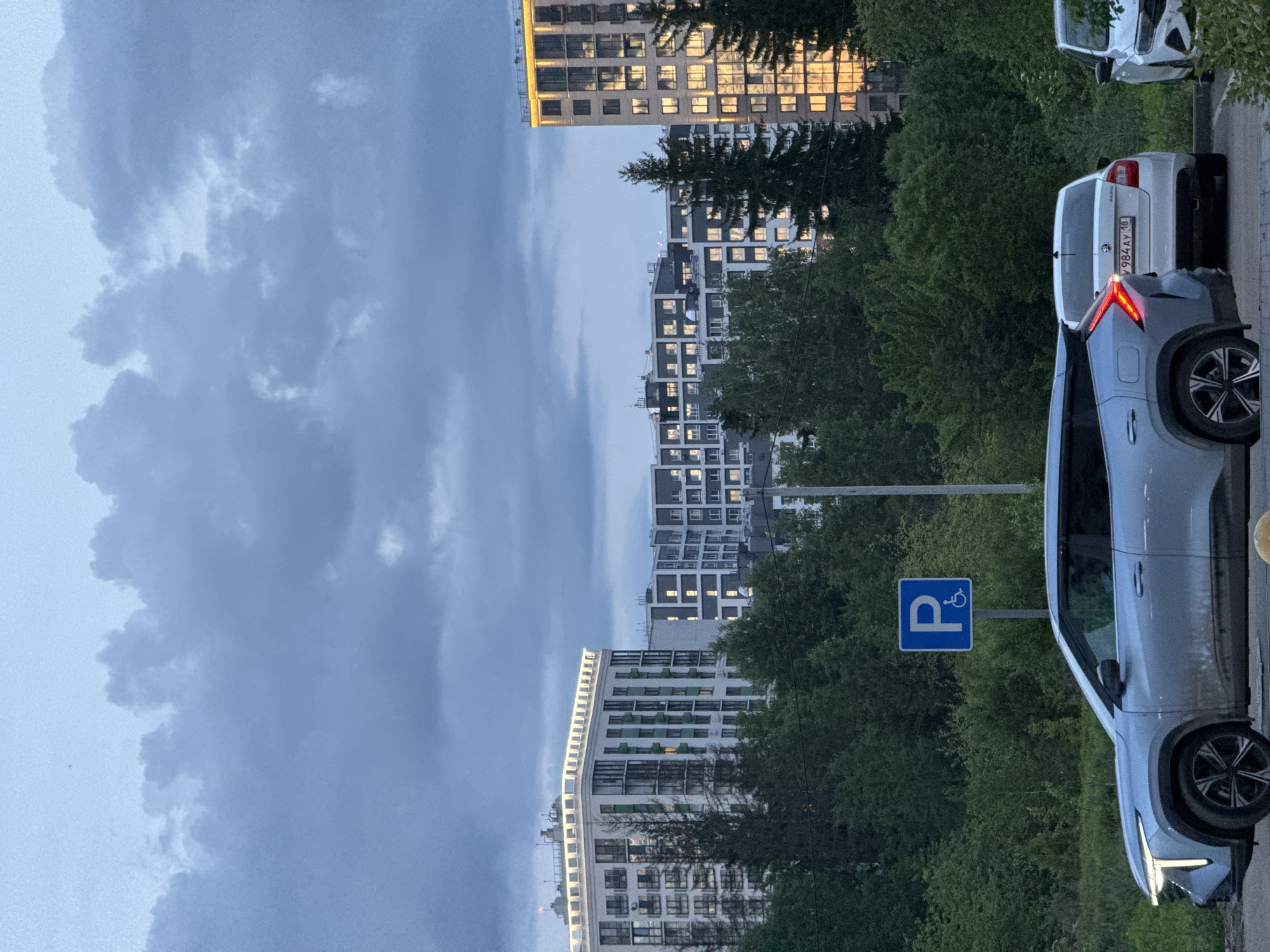 A vertical, eye-level shot depicts an urban scene likely located in Izhevsk, Russia, based on the presence of a license plate. The image features a light gray car parked near a blue parking sign with a wheelchair symbol, possibly indicating handicapped parking. Behind the car, a line of modern apartment buildings with lighted windows rises into a cloudy sky. Trees fill the space between the buildings and the parking area. Two other parked cars are visible in the background on the right. The scene seems to be captured at dusk or in overcast conditions, as indicated by the subdued light.
