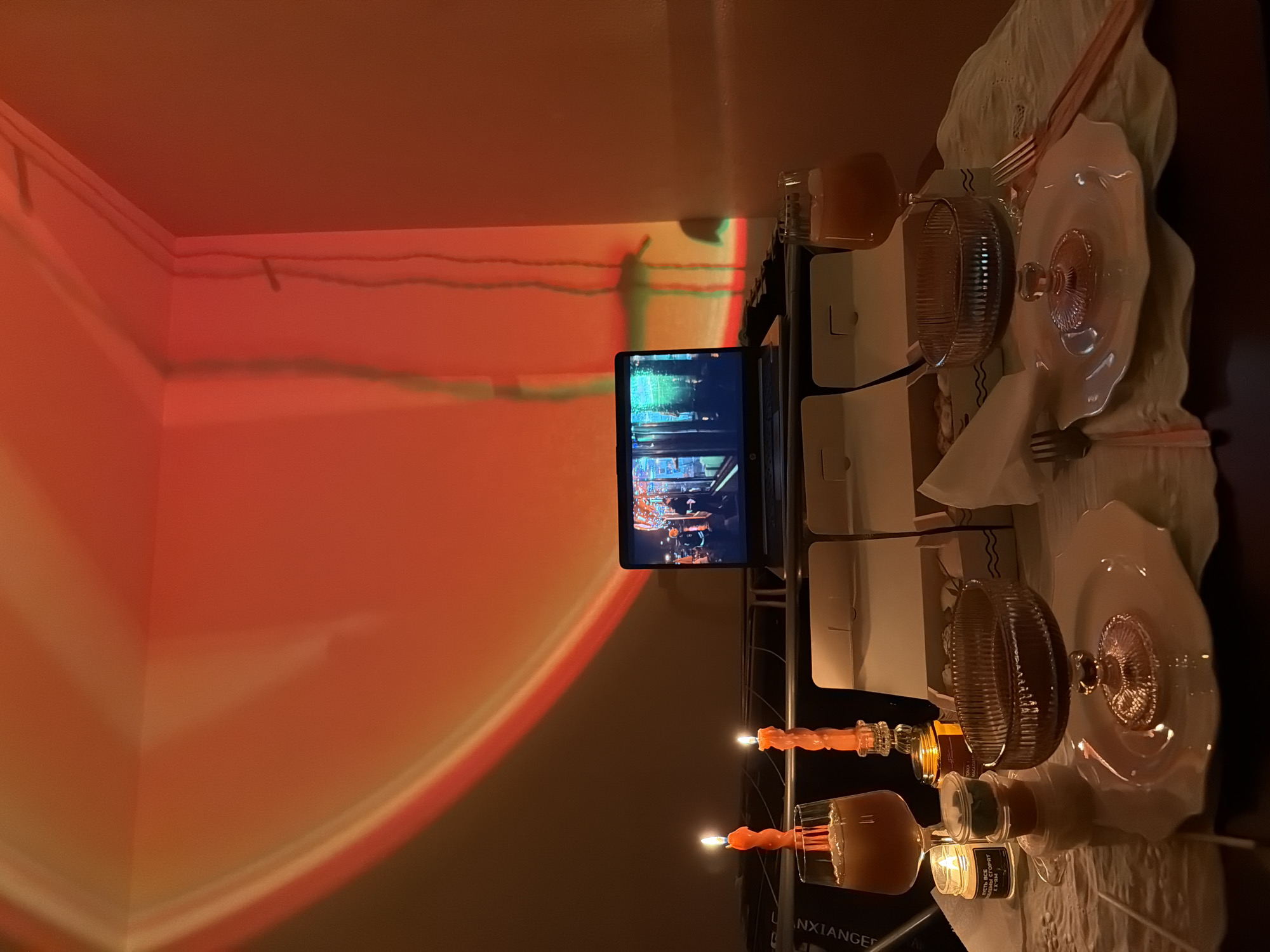 Here is a description of the image:
The image shows a set dining table in a room with warm, reddish lighting. The table is adorned with plates, glasses, silverware, and cloth napkins. Candles are placed on the table. A laptop or tablet displaying a video is also present.
There is no indication in the image that would allow me to determine the location as Izhevsk, Russia, or any other specific place.