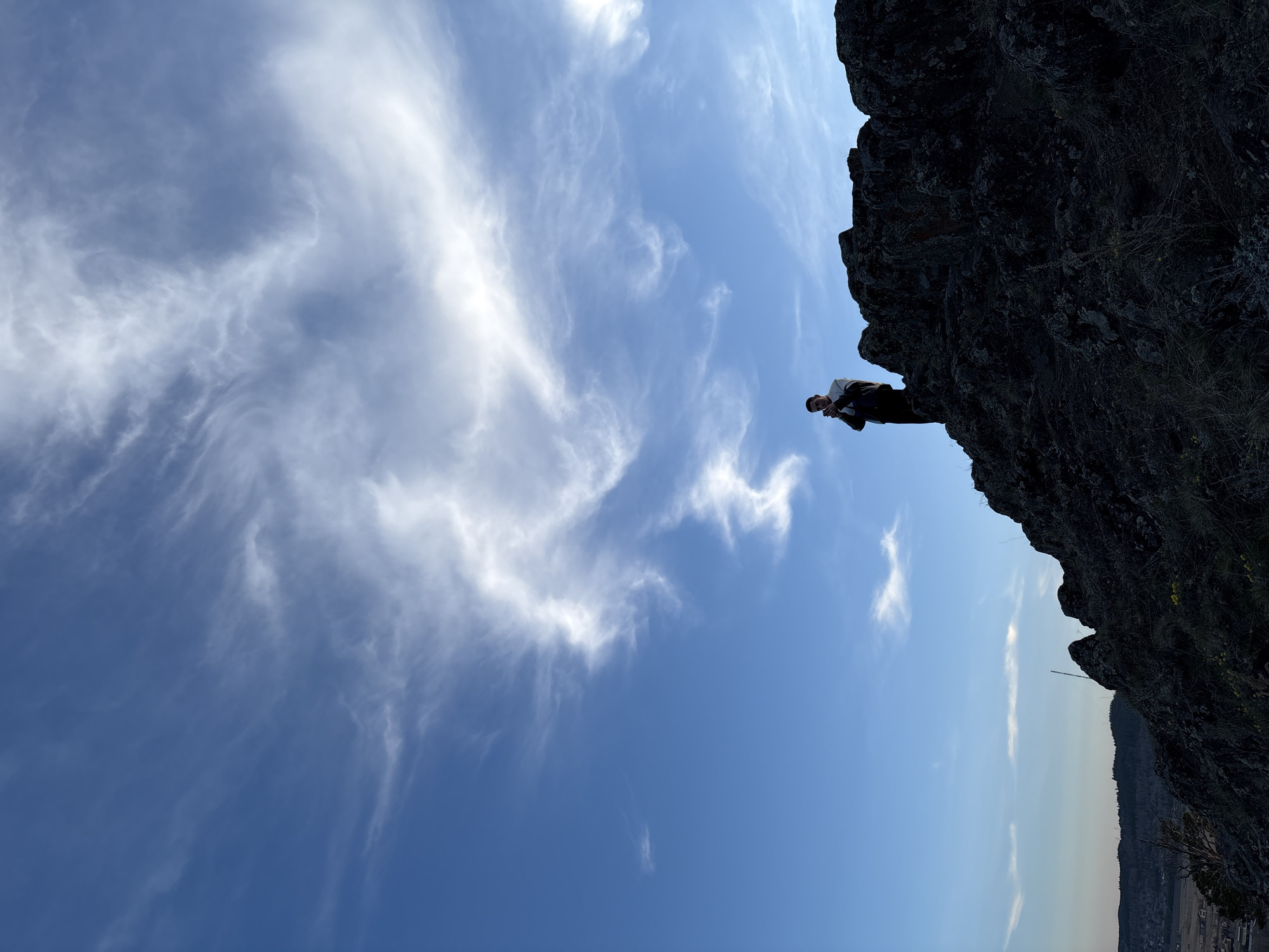 The image shows a low-angle shot of a man standing on a rocky precipice. The sky behind him is blue with wispy white clouds. Below the rock formation is a view of rolling hills and a city in the distance. Based on the location in Невон, Russia, it can be assumed this is a natural setting. The man is dressed in dark clothing and appears to be holding something in his hands.