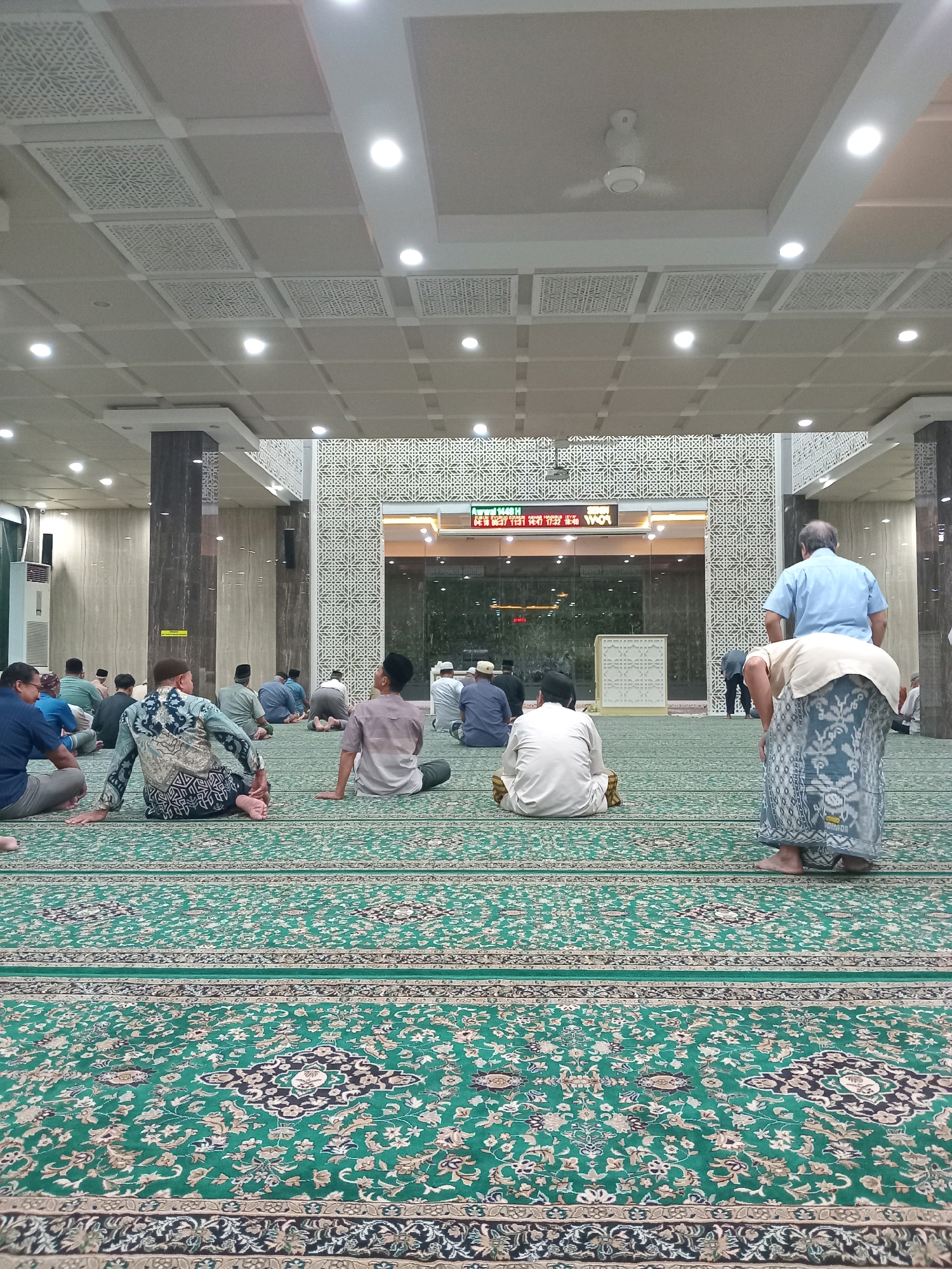 This image depicts the interior of a mosque in Randuagung, Indonesia. Numerous men are seated and kneeling on a large, green, intricately patterned carpet, facing towards the front of the prayer hall. The architecture features modern elements such as a dropped ceiling with decorative panels and recessed lighting, as well as large pillars. A digital display board is visible at the far end, showing prayer times. One man in the foreground is standing, bent over, with another man in front of him seated. The atmosphere appears to be one of prayer or religious gathering.
