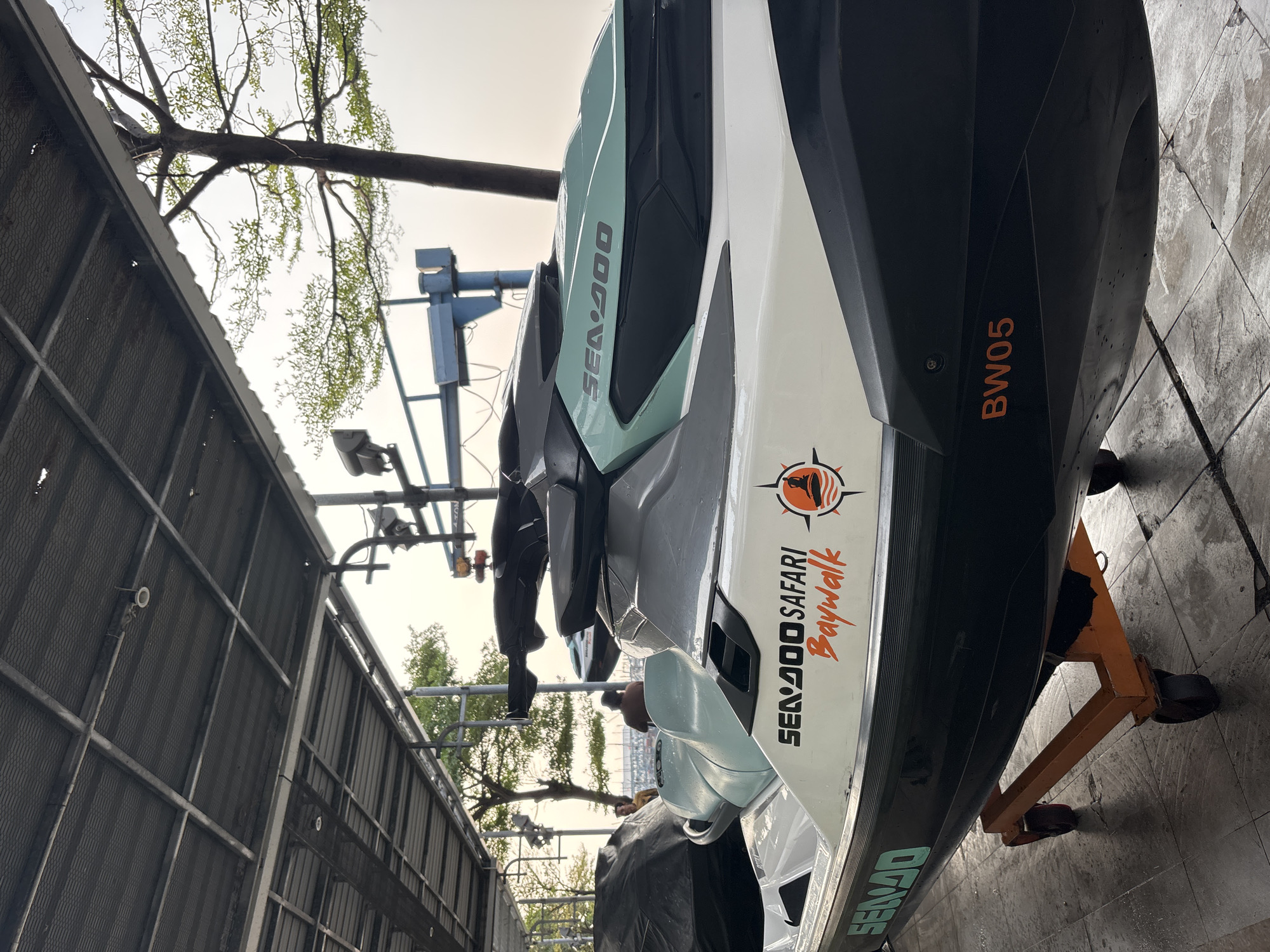 The image shows a Sea-Doo personal watercraft, possibly a jet ski, tilted on an orange stand. The watercraft has a white and black body with teal accents. Visible text includes "SEA-DOO" in teal, "SEA-DOO SAFARI Baywalk" in black and orange with an orange circular logo depicting a sunset over water, and "BW05" in orange. The background features a metal structure, possibly a warehouse or a workshop, with some trees and foliage visible against a bright, overcast sky. The lighting suggests it is daytime, likely morning or afternoon, with diffused light indicating cloudy weather. The ground appears to be concrete and slightly wet. There are no people visible in the image. The scene is set in an outdoor or semi-outdoor area, possibly a boat yard or a storage facility, given the presence of the stand and the industrial-looking structure. The overall impression is one of a functional or storage setting for the watercraft.