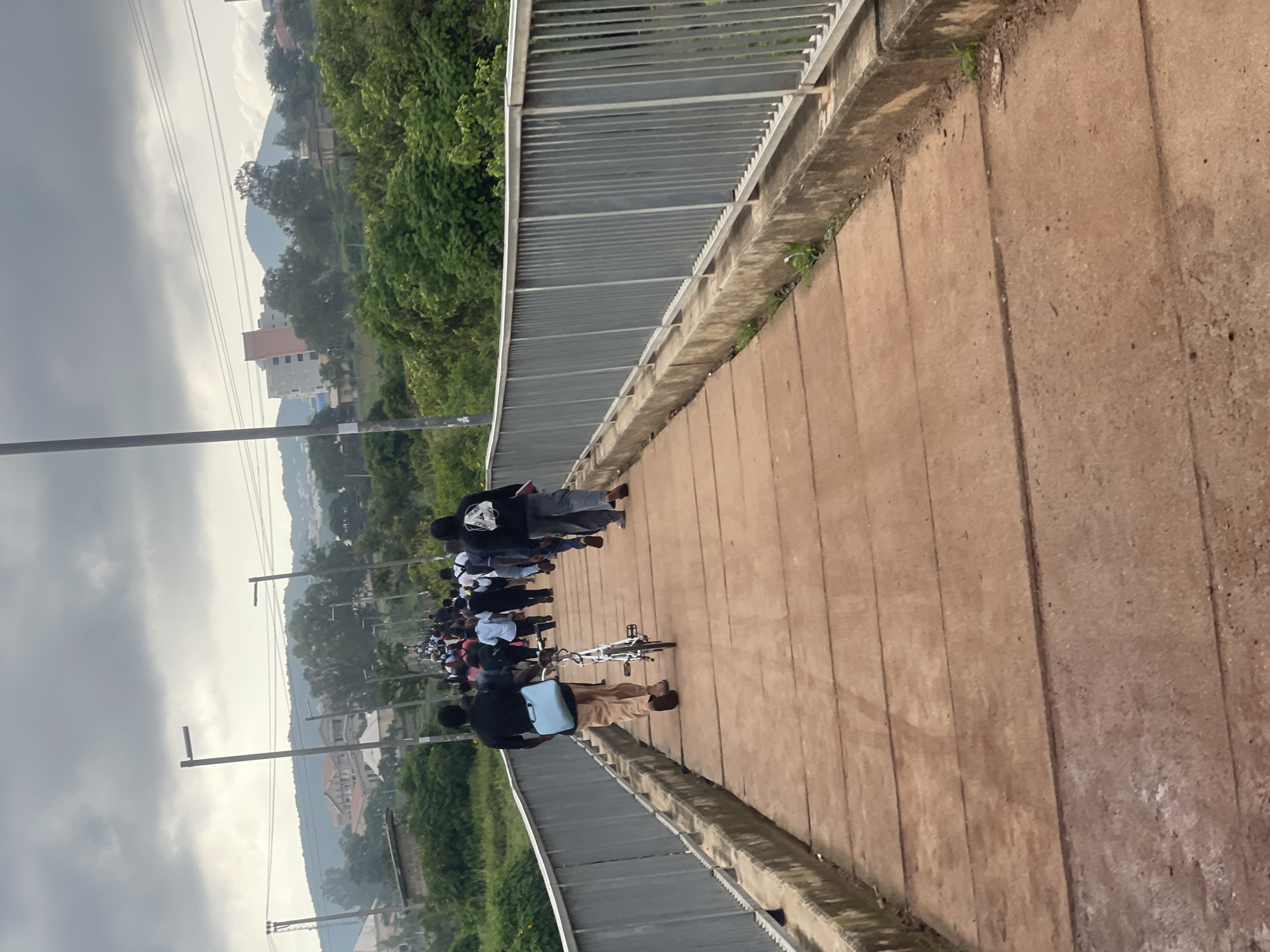 A group of people are walking on a bridge over a moderately cloudy day. In the foreground, a man in a black shirt and tan pants walks away from the camera, pulling a white bicycle behind him. He has a light blue bag slung over his shoulder. Behind him, a crowd of people, mostly wearing dark clothing, walks in the same direction. The bridge has a concrete walkway and a metal railing on the right side. In the background, there are green trees and buildings on a hillside. The scene appears to be in an urban or suburban area. The location is identified as Rafin Par, Nigeria, suggesting this is a local bridge within that city.