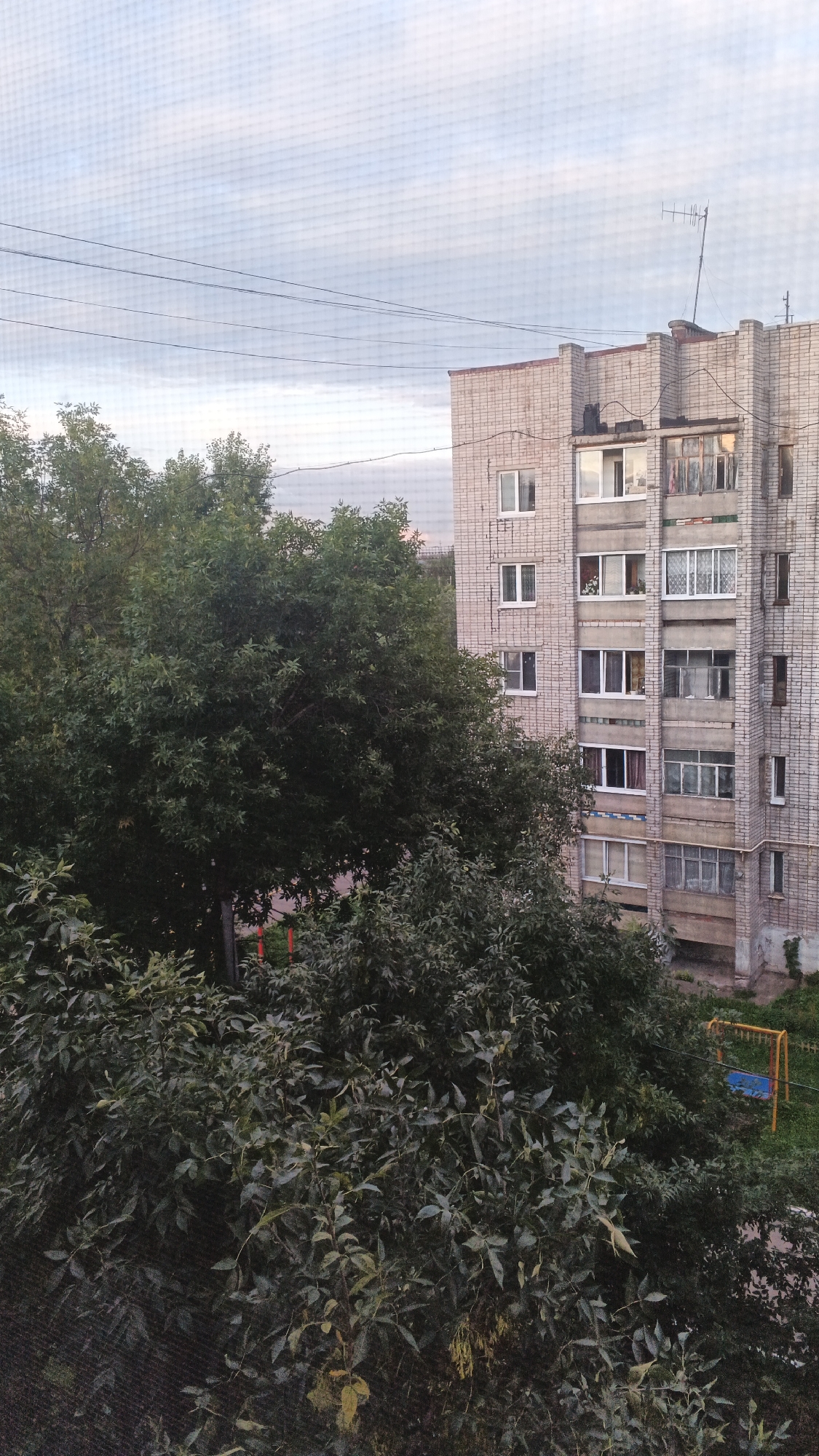 The image shows an apartment building in Izhevsk, Russia, with trees in the foreground. The building appears to be made of light-colored bricks and has multiple floors with many windows. Some of the windows have balconies or enclosed sunrooms. A few windows are open, and one shows what appears to be a curtain or bedding. There is an antenna on the roof of the building. In front of the building, there are dense green trees, some of which are tall and leafy, obscuring parts of the lower floors. In the lower right corner of the image, a yellow swing set with a blue seat is visible in a grassy area next to the building. The sky is overcast with pale clouds. Power lines are visible in the upper portion of the image.