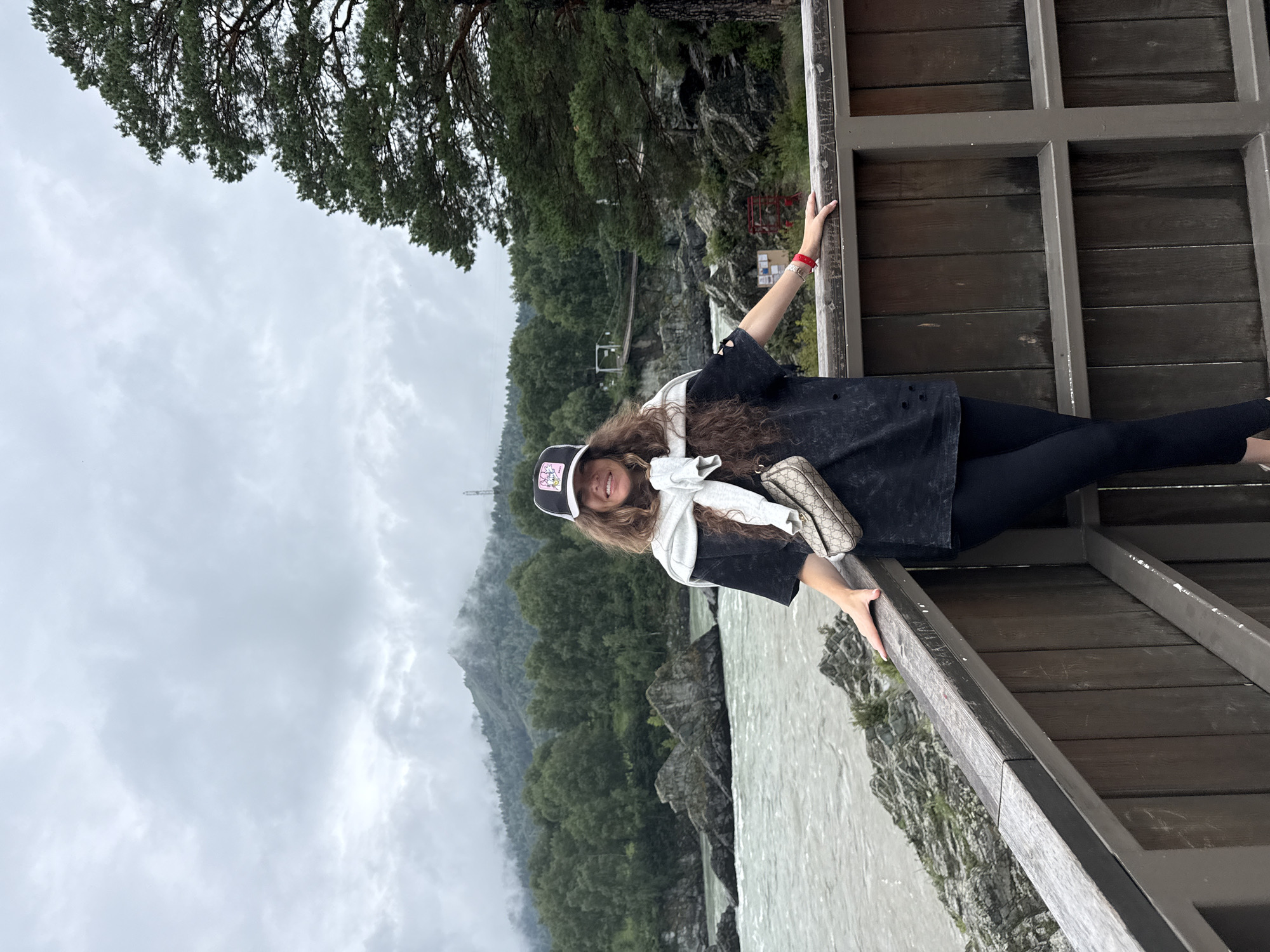 A woman stands on a wooden balcony overlooking a river and a mountainous landscape. She is wearing a black t-shirt, black leggings, a white sweater draped over her shoulders, and a black baseball cap. A beige and gold patterned purse is slung across her body. The woman is smiling and looking at the camera. The river is wide and appears to be flowing quickly, with large rocks in the water and along the banks. The background features a dense forest and mountains, with clouds obscuring the peaks. The overall mood of the image is relaxed and scenic. The location is identified as Elekmonar, Russia.