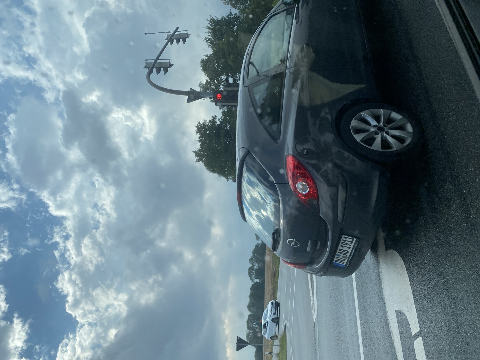 A dark gray Opel Corsa is stopped at a traffic light on a wet road. The license plate reads "DU AB 1911". A white Volkswagen Golf is visible in the distance, also on the road. The sky is overcast with dramatic clouds, and trees line the horizon. The scene appears to be an intersection, indicated by the traffic signals and road markings. This is likely a scene in Weyhe, Germany, given the license plate format.