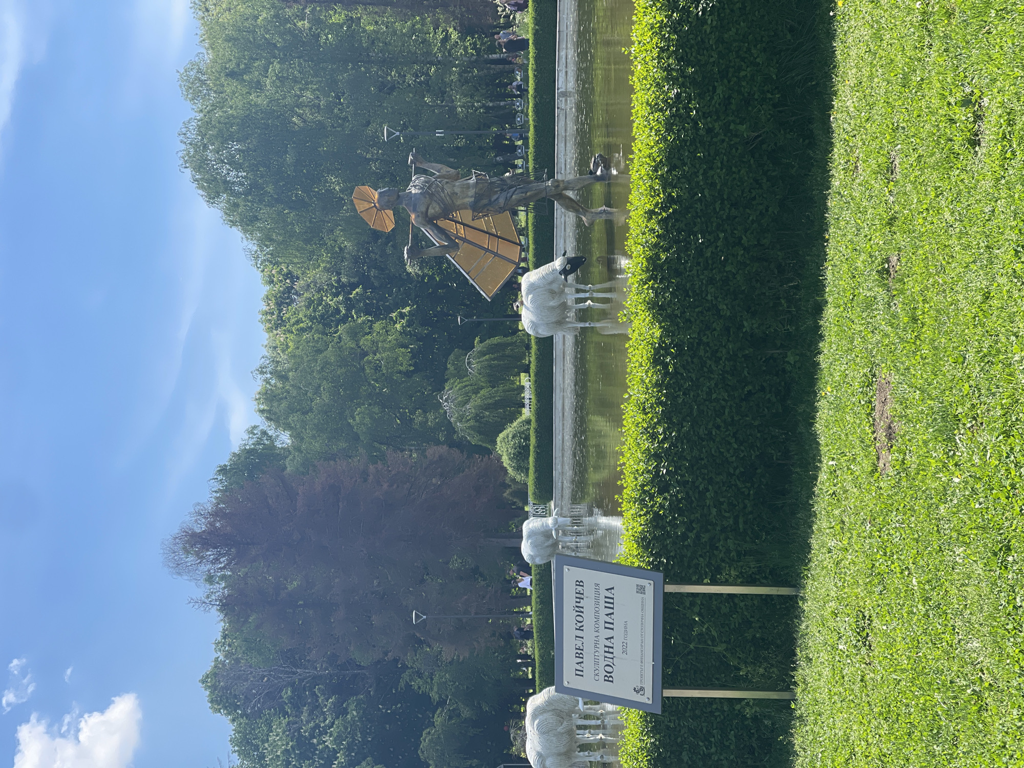 The image shows a sculpture in a pond in a park in Sofia, Bulgaria. The sculpture depicts a person with wings and a golden parasol, accompanied by white sheep. A sign next to the pond identifies the sculpture as "Pavel Koychev: Water Pasha," created in 2022. The pond is bordered by a green hedge and grassy area. Trees and a blue sky with some clouds are visible in the background.