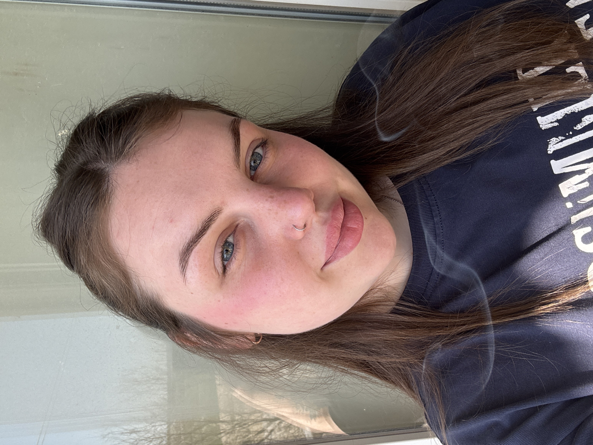 A young woman with fair skin, brown hair, and blue eyes is facing the camera. She has a nose ring in her left nostril and is wearing a dark blue t-shirt with white lettering. Smoke or a hazy effect is present near her hair and shoulder. The background appears to be a window or glass panel with an obscured outdoor view, possibly including trees. The image location cannot be determined from the information available.