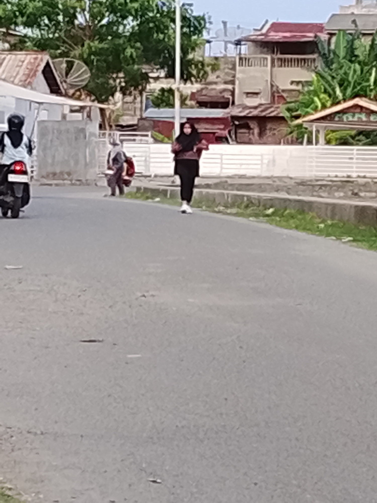 In Lhokseumawe, Indonesia, a street scene unfolds with several individuals and a parked motorcycle. On the left, a person wearing a helmet and a black hijab rides a motorcycle, facing away from the camera. To their right, another person in a grey hijab is crouched down, holding a red object. Further to the right, a person in a black hijab and black pants walks away from the camera, wearing white shoes. The background features buildings with varying roof colors, some with balconies, and dense green trees. A white railing runs along the side of the road. The road surface is asphalt, with some debris visible. The lighting suggests it is daytime.