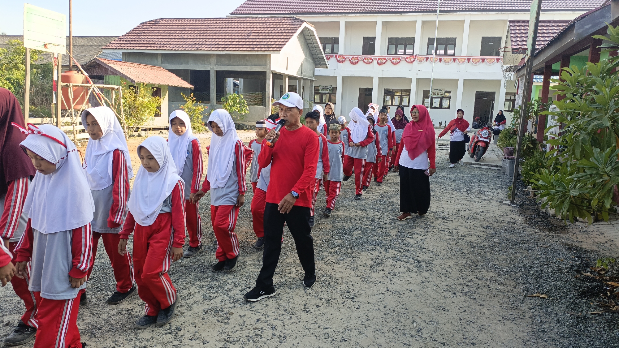 In Saring Sungai Binjai, Indonesia, a group of students in red and gray uniforms, accompanied by a man holding a microphone and wearing a red shirt and black pants, walks on a gravel path. The students, both male and female, wear white hijabs and appear to be participating in an outdoor activity. Behind them, a white building with a red-tiled roof stands, suggesting it is a school or community center. The environment is sunny with vegetation visible. A red motorcycle is parked nearby, and other individuals are seen in the background, indicating a public gathering or event.