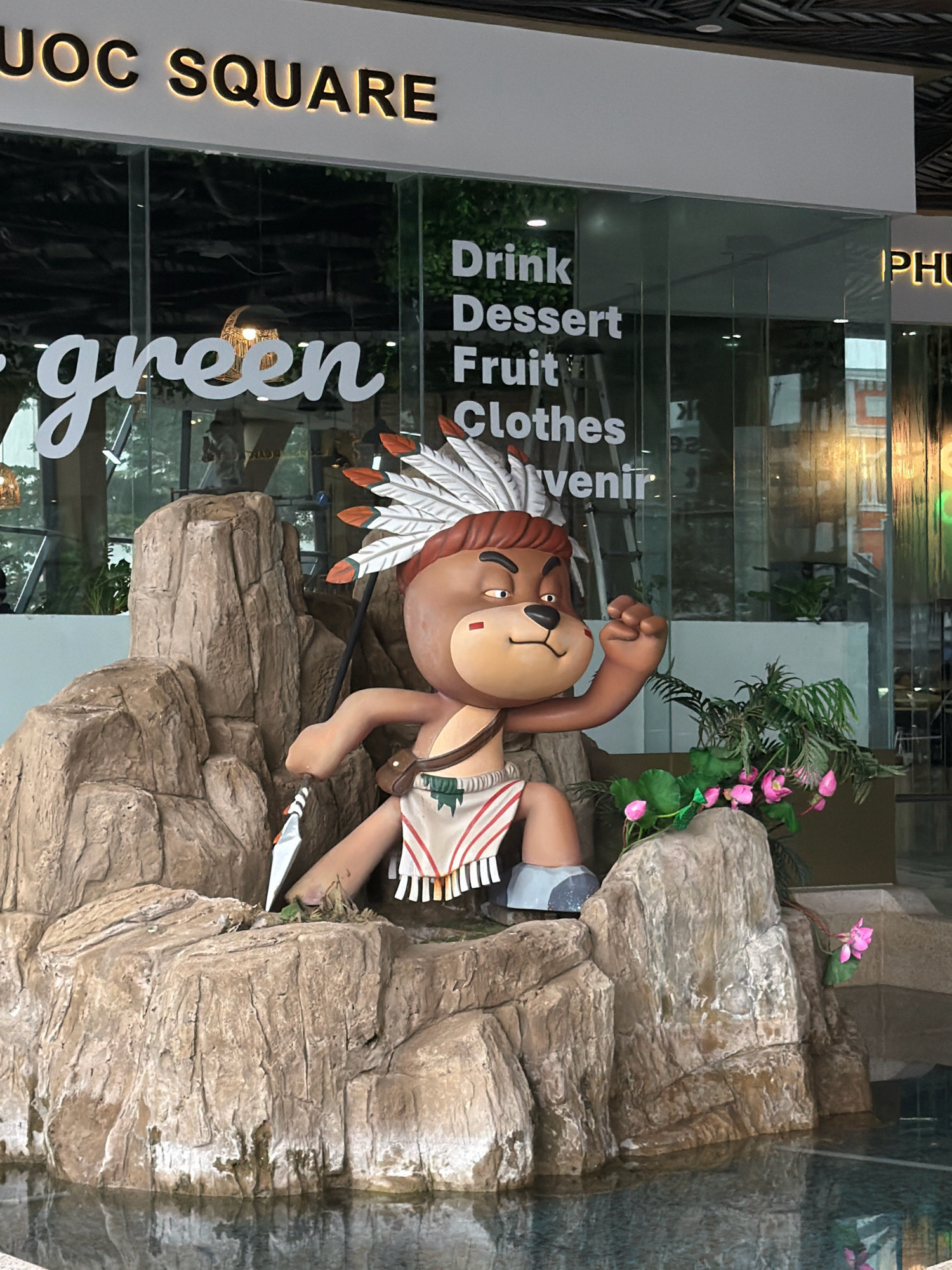 The image captures an outdoor scene in front of a building with a prominent sign that reads "UOC SQUARE". To the left of this sign, a large, white cursive script reads "green". A statue of a cartoonish bear character, dressed as a Native American with a headdress and loincloth, sits atop a rocky outcrop. The bear is holding a spear and has its fist raised. In the background, through glass windows, a store interior is visible, with signage including "Drink", "Dessert", "Fruit", "Clothes", and "Souvenir". To the right of "UOC SQUARE", a sign reads "PHU". Pink flowers and lush green foliage are situated near the base of the statue. The rocky outcrop is partially submerged in a body of water, reflecting the surroundings. The overall impression is that of an entrance to a commercial establishment, likely a shopping area or mall, in a city.