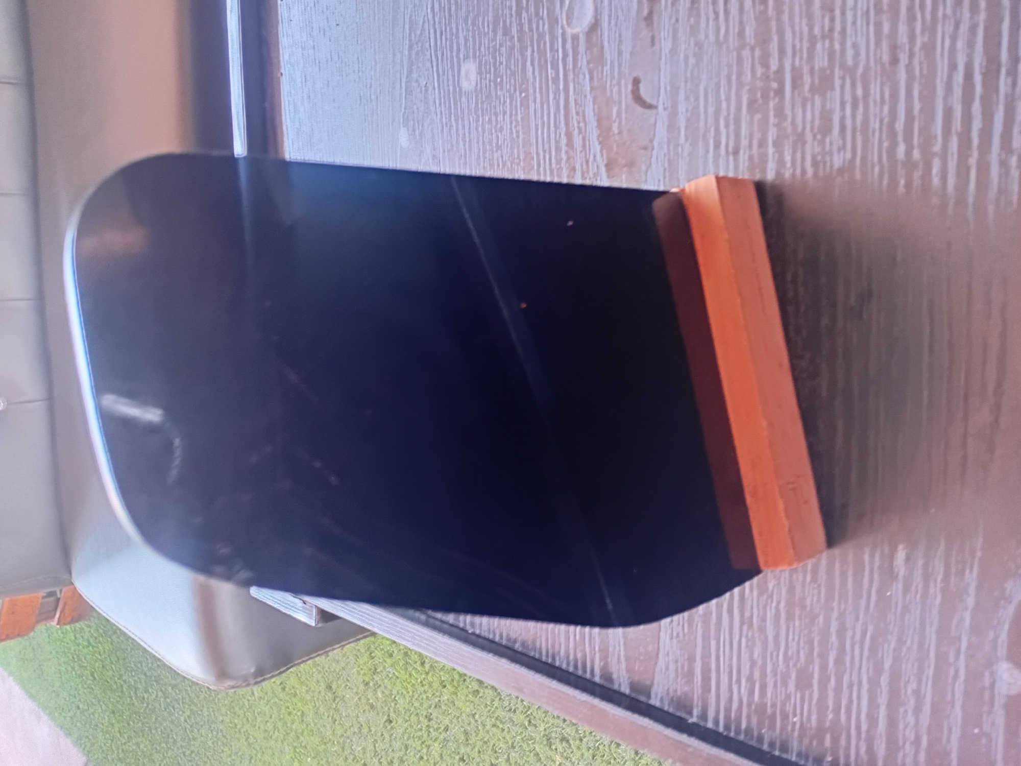 A reflective black object with rounded top edges stands upright on a light brown wooden base. The object is positioned on a dark brown wooden table with a horizontal grain pattern. In the background, a portion of a dark gray couch is visible, with a green, grassy area to the left of the couch, suggesting an outdoor setting. The scene is indoors, as indicated by the indoor furniture and table. The location is identified as Howayi, Nigeria.