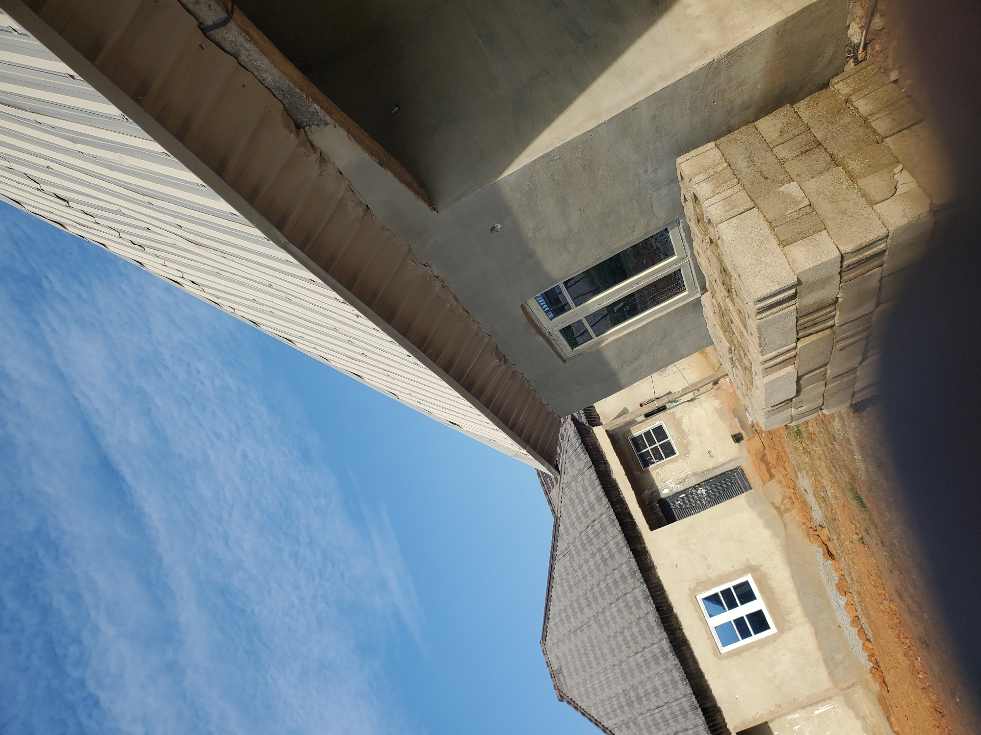 This forensic image, taken in the city of Winters, United States of America, depicts a construction or recently completed residential area under a clear blue sky with sparse, wispy white clouds. In the immediate foreground, a stack of light grey concrete blocks sits on reddish-brown exposed earth. Adjacent and angled sharply on the right, a building with an unfinished, light grey stucco or concrete exterior features a prominent light beige corrugated metal roof and a single, white-framed window with reflective glass. To the left and further back, a second building, also appearing unfinished, presents a light tan exterior, a dark grey multi-gabled roof, two white-framed windows, and a partially open dark-colored entry door. The ground around both structures is predominantly reddish-brown dirt, characteristic of a building site, with minor ground debris. No individuals are present, and no active work is visibly occurring.