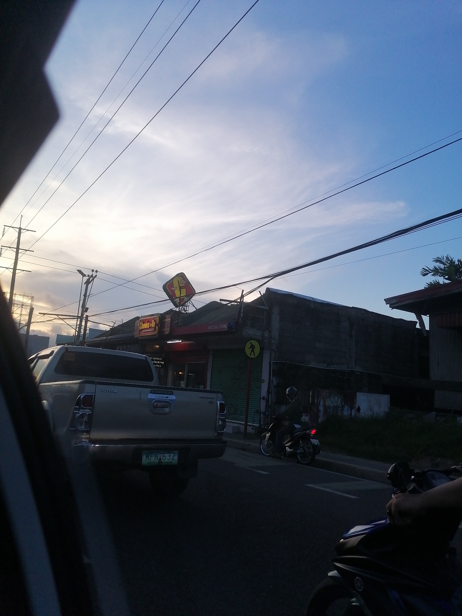 The image captures a street scene in Matina, Davao City, Philippines, during what appears to be late afternoon or early evening. The sky, occupying the upper half, is light blue with scattered thin white clouds, suggesting a bright but possibly setting sun towards the left horizon. Multiple black utility lines traverse the sky horizontally and diagonally.
In the midground, a silver/grey Toyota Hilux pickup truck, model "VIGO G," is prominently featured, facing away from the viewer. Its license plate, "MFN 632," is clearly visible. The vehicle appears to be in traffic, possibly stopped or moving slowly.
On the left side of the road, behind the Toyota, a commercial building features a large, distinct yellow and red sign for "Chooks-to-Go." Below it, a smaller red sign displays "MATINA DAVAO," confirming the location. Another small sign below the main one reads "tattoo piercing coffee." The building's front is secured by green metal rolling shutters. A utility pole with multiple wires stands near the building, and a yellow circular pedestrian crossing sign is visible on a pole nearby.
On the right side of the road, a dark-clad individual wearing a helmet is seated on a motorcycle that is stopped near the curb. Further into the foreground on the far right, a partial view of another motorcycle and a person's hand holding handlebars are visible. The road surface is asphalt with white dashed markings. A concrete wall and some vegetation are visible on the right side, behind the motorcycles, with a building featuring a reddish roof further in the background. The scene is partially framed on the far left by a dark, irregular shape, consistent with the interior of a vehicle from which the photograph was taken.