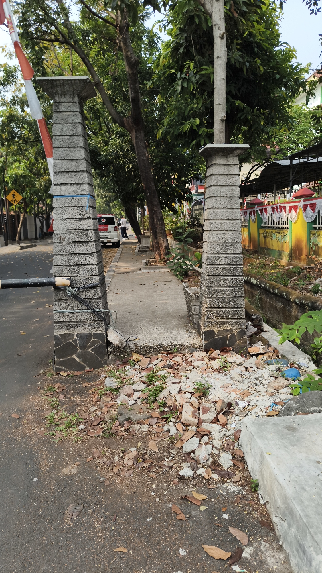 The image depicts a paved walkway with two stone pillars on either side, forming an entrance. Debris, including broken bricks and dried leaves, is scattered on the ground in front of the pillars. A black metal barrier extends from the left pillar across the walkway. A white van with a red cross emblem is parked on the street to the left of the walkway, and a person is standing near it. Trees line the walkway, and a decorative fence with red and white trim is visible in the background. The scene appears to be in a residential area of South Jakarta, Indonesia, indicated by the architectural style and the Indonesian language on a yellow traffic sign in the background.