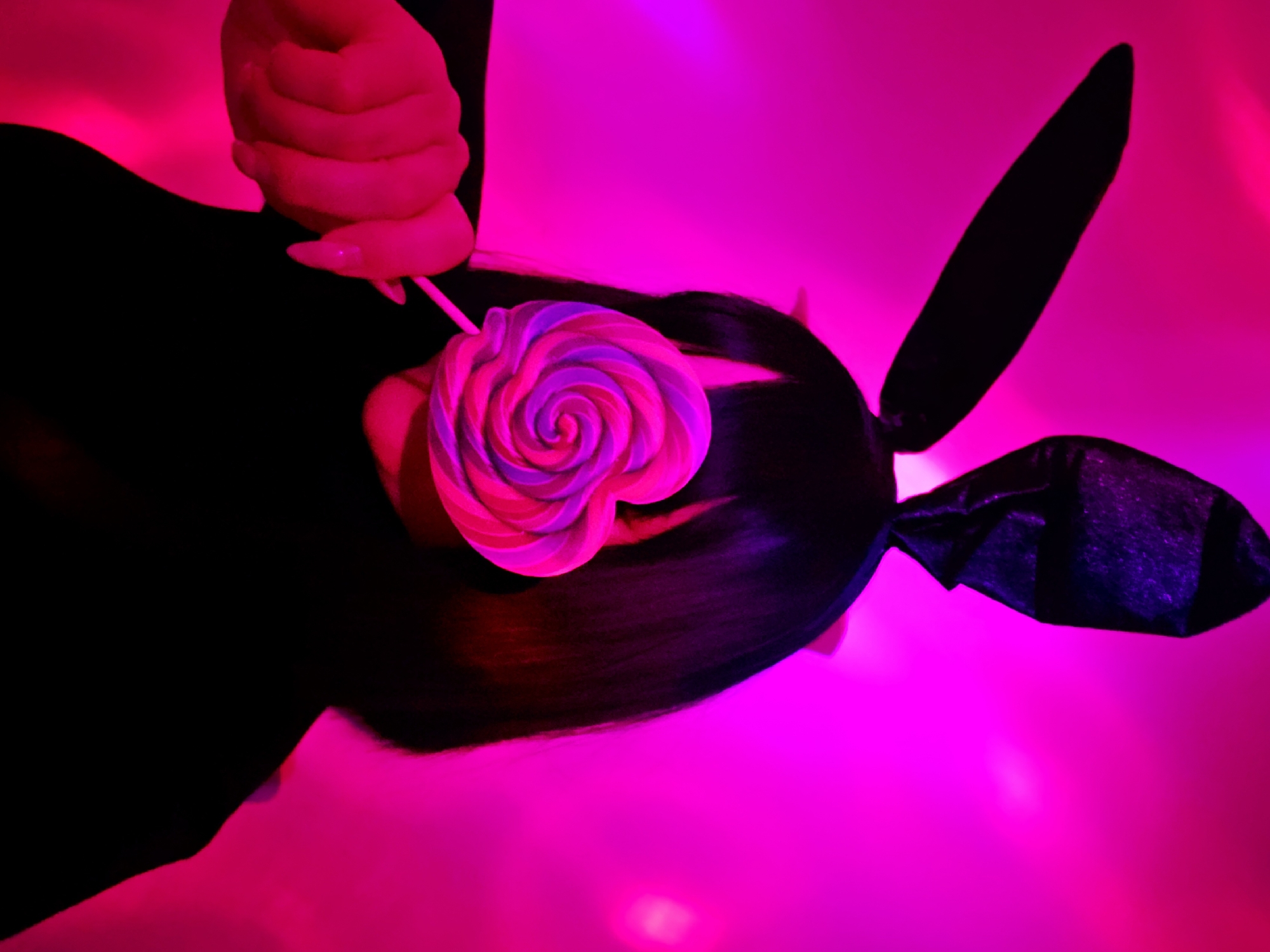 A view from above, illuminated with magenta lighting, depicts a person wearing black bunny ears and holding a lollipop. The individual's dark hair is visible, and a hand is positioned near the lollipop, held close to their face. The background has a distinct magenta hue.