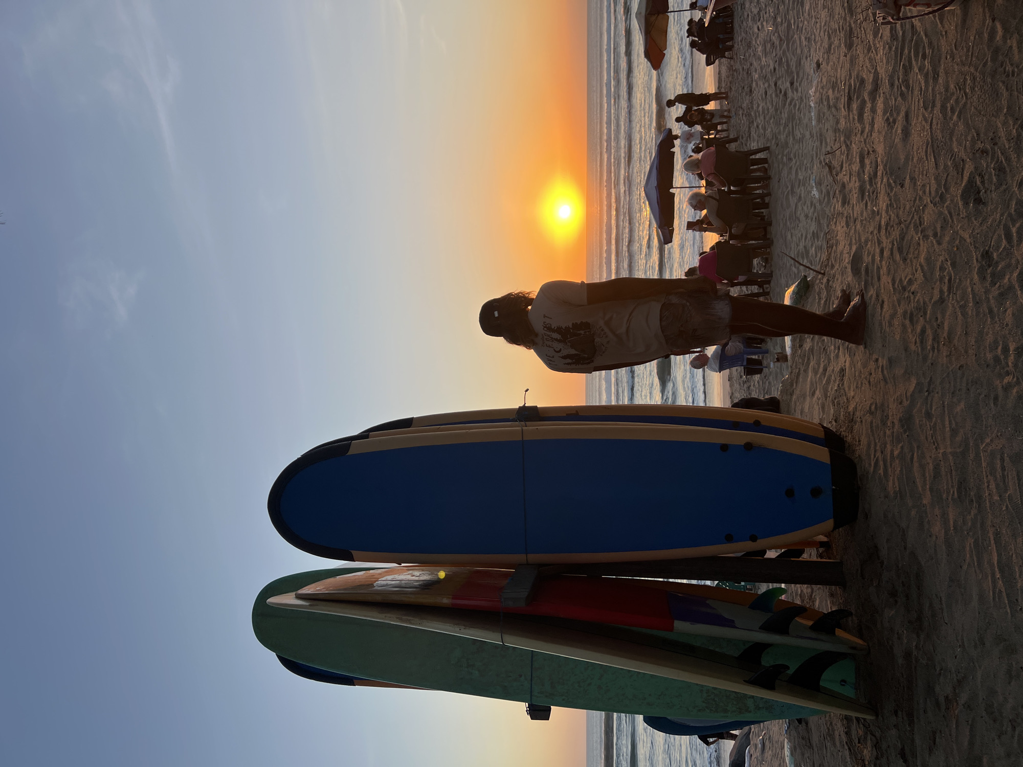 At sunset on a beach in Kuta, Indonesia, a man stands facing the ocean, his silhouette framed by the vibrant orange sky. In the foreground, a rack of surfboards stands tall, their colorful designs hinting at the day's activities. The sand is dotted with people enjoying the view, seated at tables under umbrellas, their forms softened by the fading light. The waves of the Indian Ocean gently roll onto the shore, reflecting the warm glow of the setting sun. The scene is one of peaceful closure to a day, a moment of shared appreciation for nature's beauty.