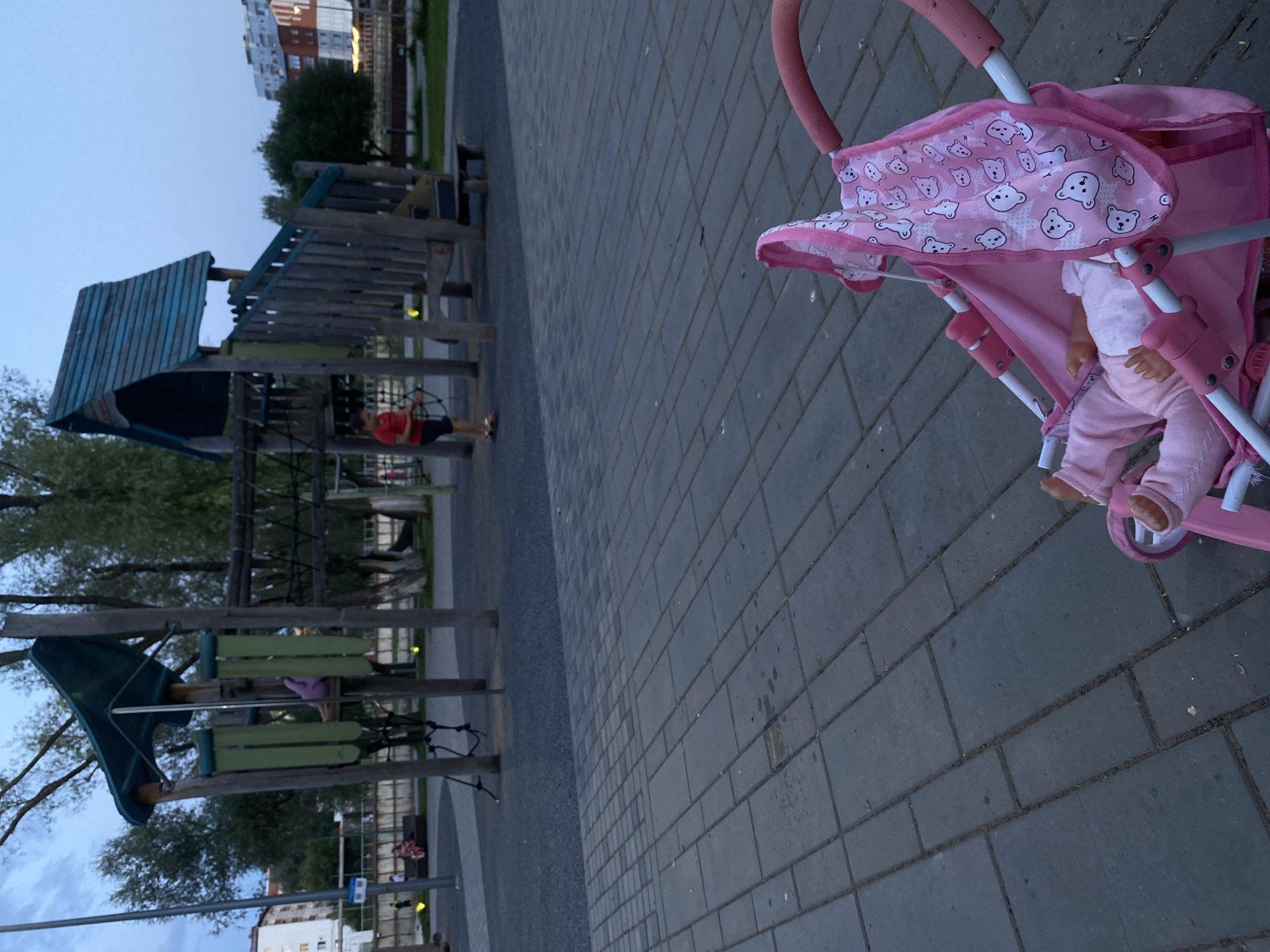 In Votkinsk, Russia, a pink doll stroller with a doll wearing pink clothes lies on a paved walkway in a playground area. The stroller is in the foreground, with its canopy adorned with a pattern of white bears. In the background, a wooden playground structure with slides and climbing elements stands under a twilight sky. A young boy in a red shirt and dark shorts is visible near the playground, appearing to interact with a wheel-like component. Other children and adults are scattered throughout the playground. The environment appears to be a public park or recreational area, with trees and buildings visible in the distance.