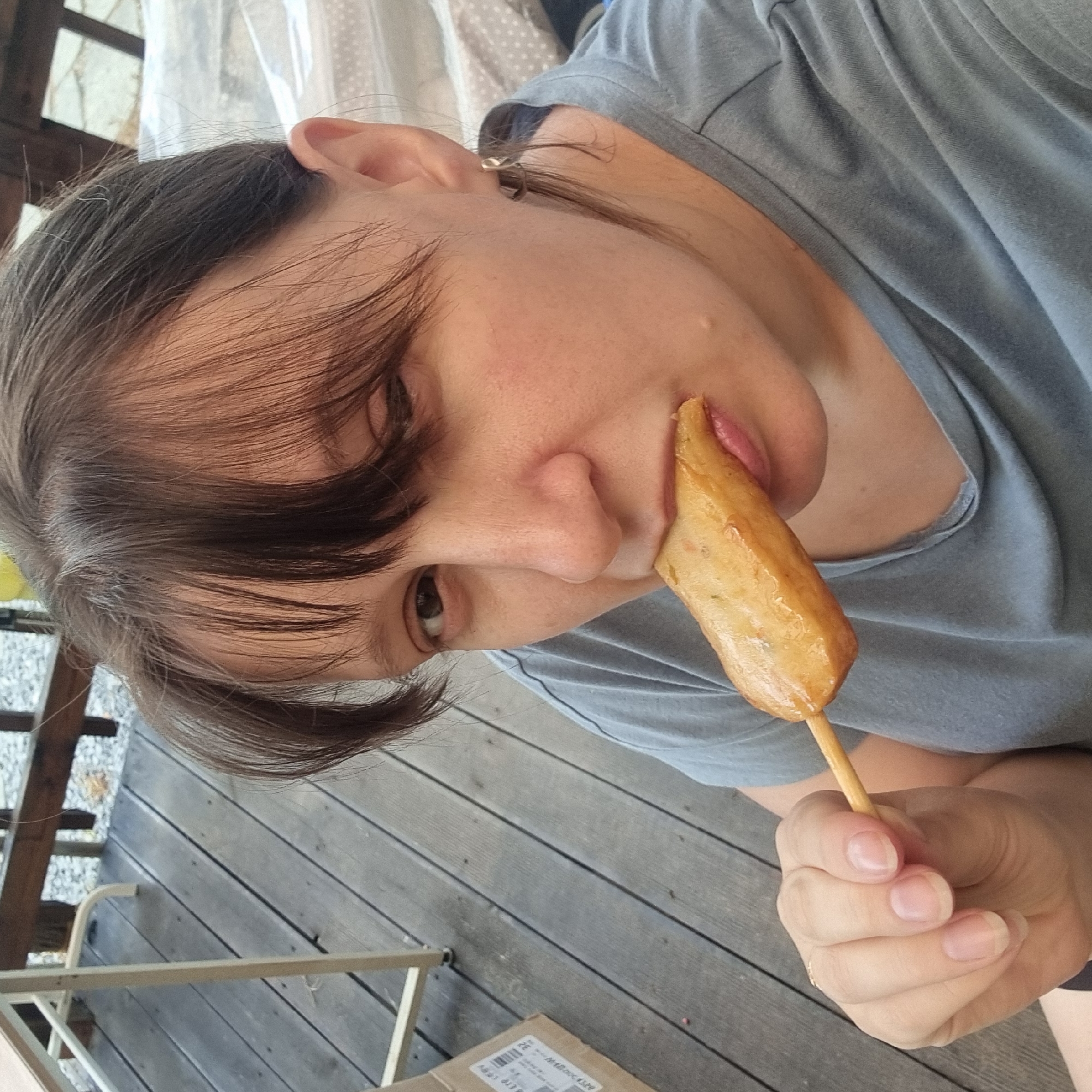 A woman is eating a piece of food on a stick outdoors. She is wearing a gray t-shirt and has dark brown hair. The food is golden brown and appears to be fish cake. She is holding the stick with her left hand. The background is a wooden deck, and a table and chairs are visible. There is a cardboard box on the deck to the left. The environment suggests a casual outdoor setting, potentially a restaurant or a home patio. The image was taken in Dunnae-myeon, South Korea.