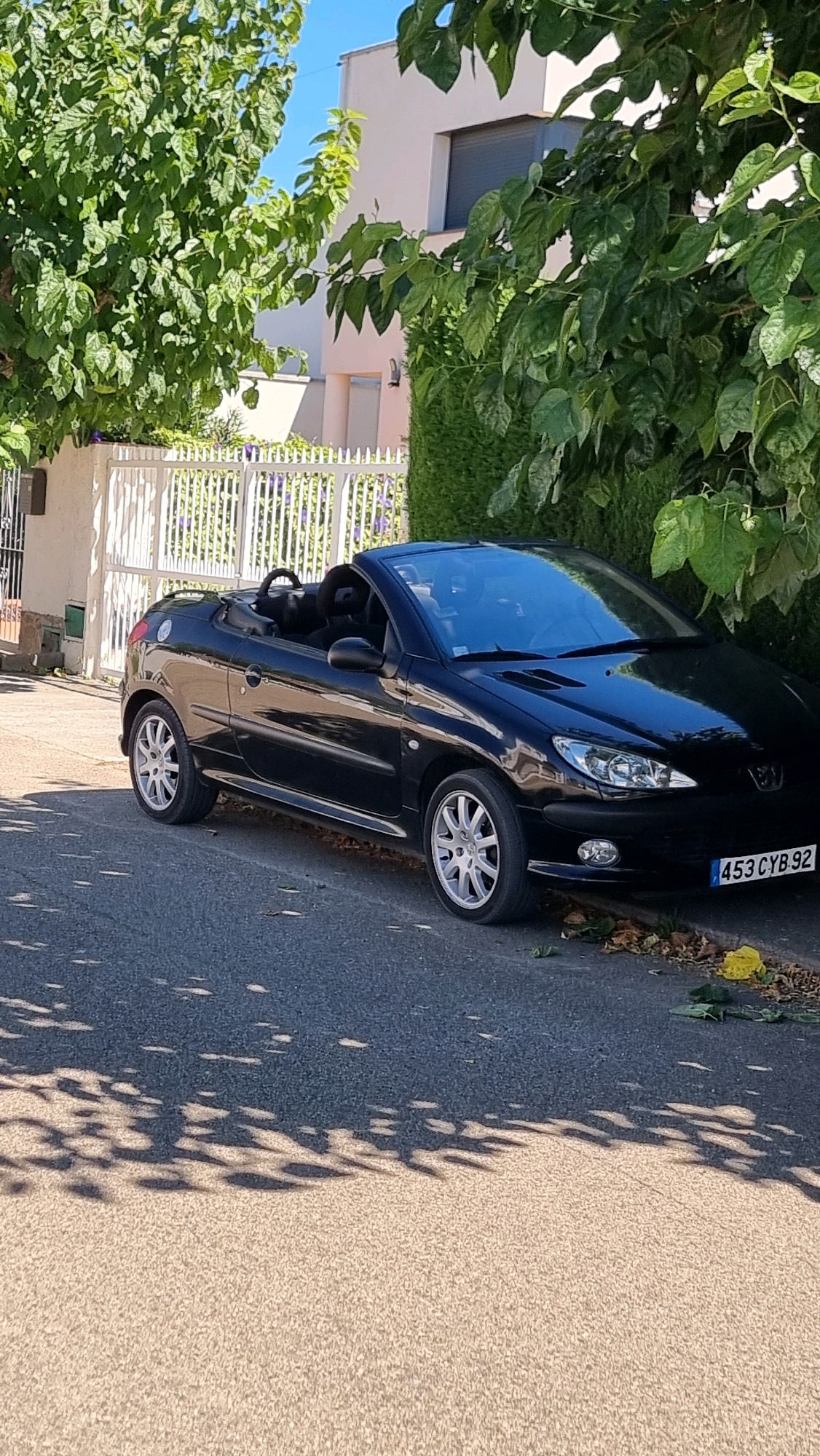 A black convertible Peugeot 206 is parked on a paved road in front of a white picket fence and a modern house. The car's top is down, revealing the black interior and seats. The license plate of the car reads "453 CYB 92". Lush green trees and bushes surround the scene, casting dappled shadows on the road and the car. The sky is clear and blue. The environment suggests a residential area, likely in a sunny climate. The city of Castelló d'Empúries, Spain, is known for its well-preserved medieval quarter and its location near the Mediterranean coast, offering a picturesque backdrop for such a scene.