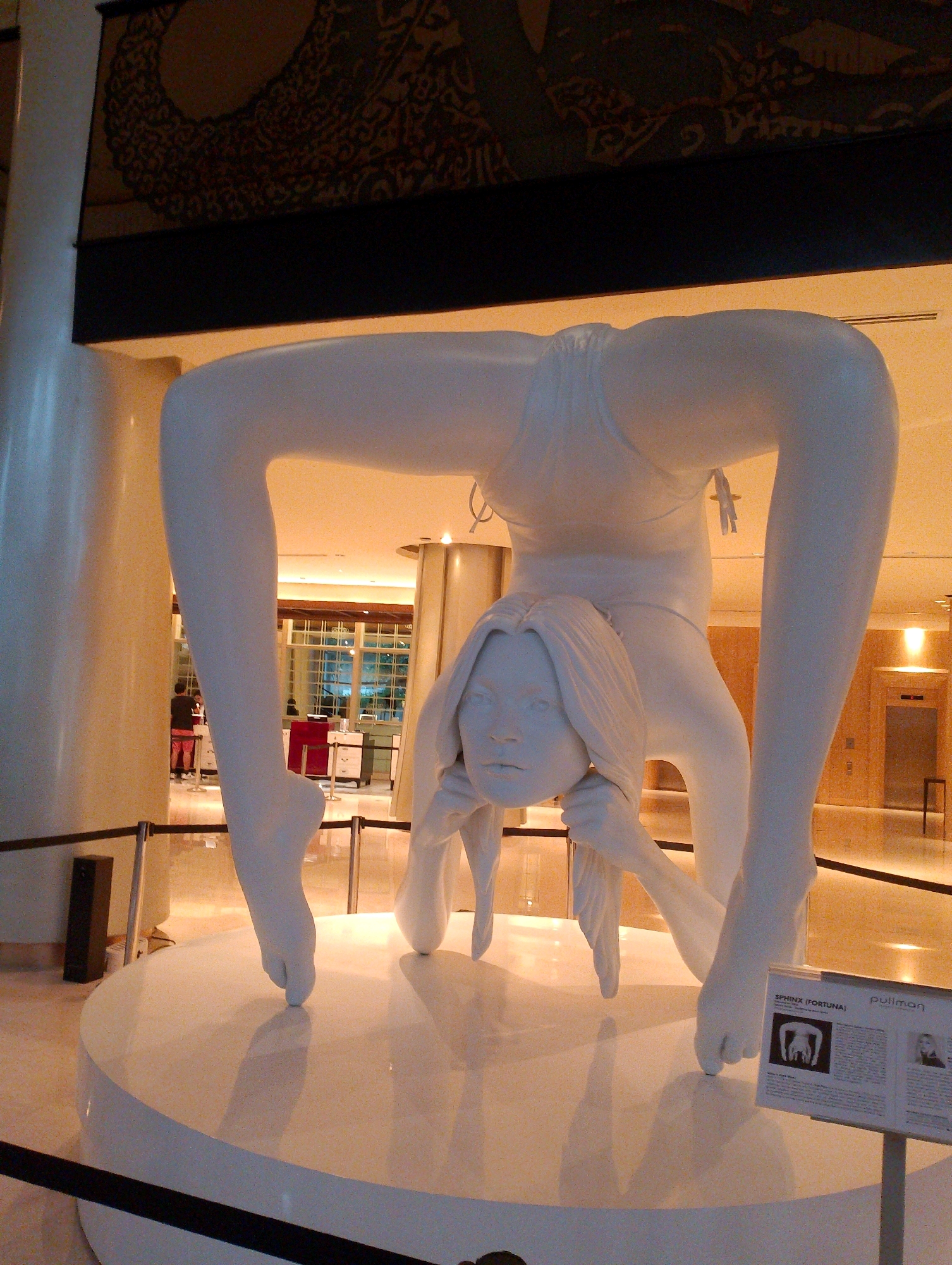 The image shows a large, white, abstract sculpture of a woman in a contorted yoga pose, resembling a crab walk or a wheel pose. The sculpture is made of a smooth, matte material and appears to be hollow. The woman's legs are bent and extended upwards, forming the main structure of the piece, while her head is inverted and rests on her hands, which are positioned on a white circular pedestal. The sculpture's body is positioned above the head, creating a sense of fluidity and dynamism.
The sculpture is located in an indoor public space with a high ceiling and warm, ambient lighting. In the background, there are large windows with horizontal blinds, revealing a glimpse of an urban environment. There are also tall, cylindrical pillars and decorative elements on the wall, suggesting a modern and elegant interior design. A sign with text and images is visible in the lower right corner of the image. The sign includes the words "SPHINX (FORTUNA)" and "pullman", indicating the name of the artwork and possibly the hotel or exhibition it is part of.
The sculpture is the central focus of the image, occupying most of the frame. The lighting casts soft shadows on the white pedestal, highlighting the curves and contours of the sculpture. The overall impression is one of artistic expression and aesthetic appeal within a sophisticated setting. The cropped image hints at the sculpture's intricate details, such as the texture of the hair and the delicate features of the face. The sculpture is positioned in a way that emphasizes its flexibility and balance. The environment appears to be a hotel lobby or a similar public space in Central Jakarta, Indonesia, given the presence of the Pullman branding. There are no people interacting directly with the sculpture in the visible frame.
