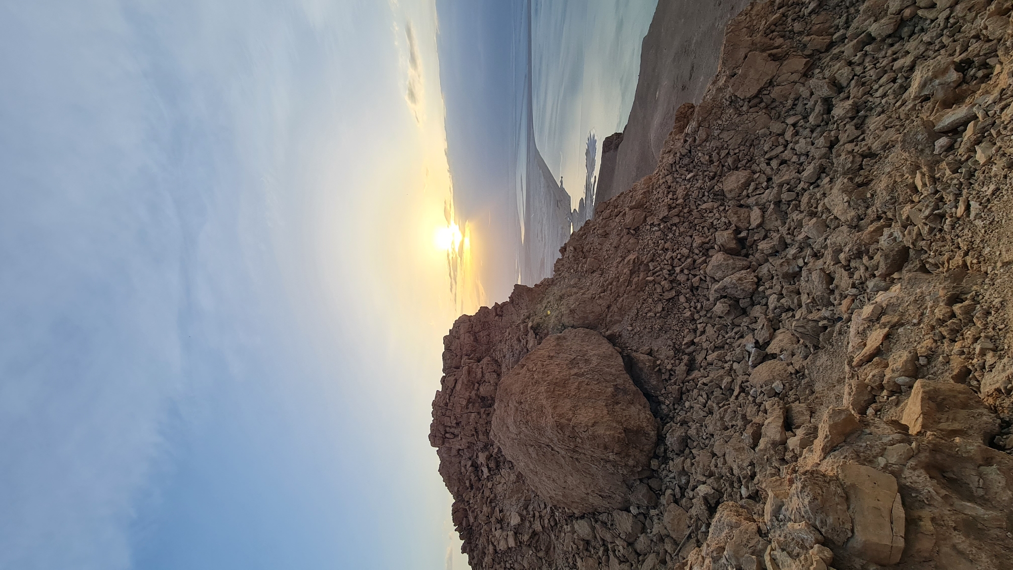 The image shows a rocky hillside at an angle, with a large boulder prominent in the foreground. Smaller rocks and earth cover the slope. Beyond the hillside is a view of a body of water under a bright, setting or rising sun, with a shoreline visible in the distance. The sky is blue with some cloud cover. This is the city of Tamar Regional Council, Israel.