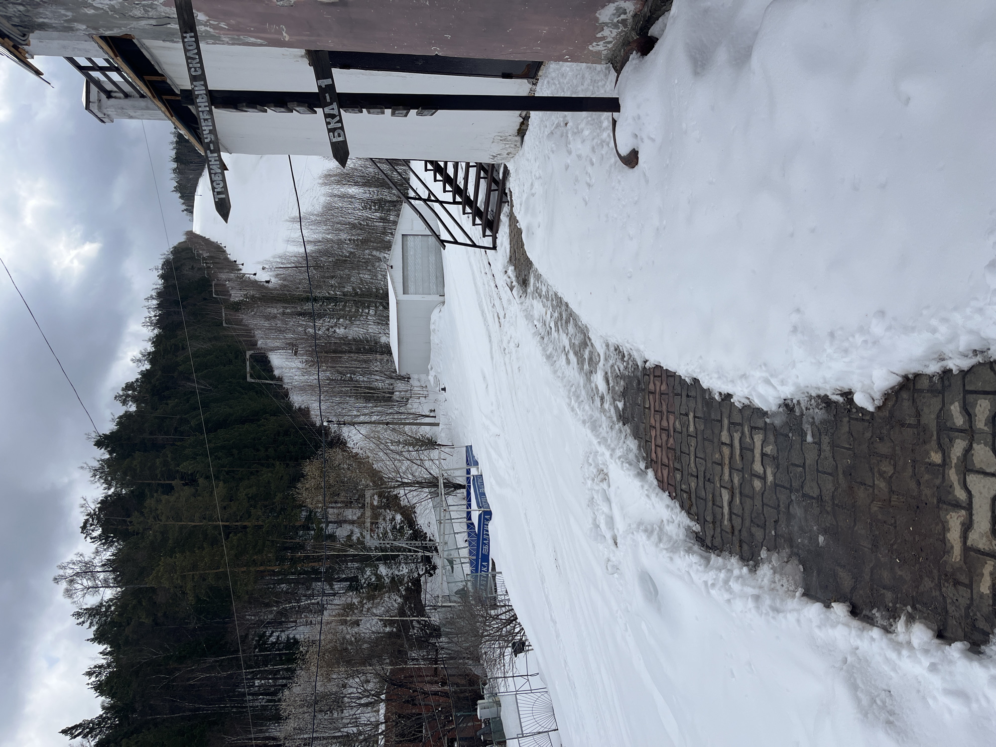 The image shows a snowy landscape, possibly a ski resort or mountainous area in Unknown. There's a building to the right with signs pointing towards "ТЮБИНГ- УЧЕБНЫЙ СКЛОН" and "ВКА-1". A snow-covered path leads from the building, with some brickwork visible. The background features a forest and a cloudy sky. A ski lift structure is present in the distance.