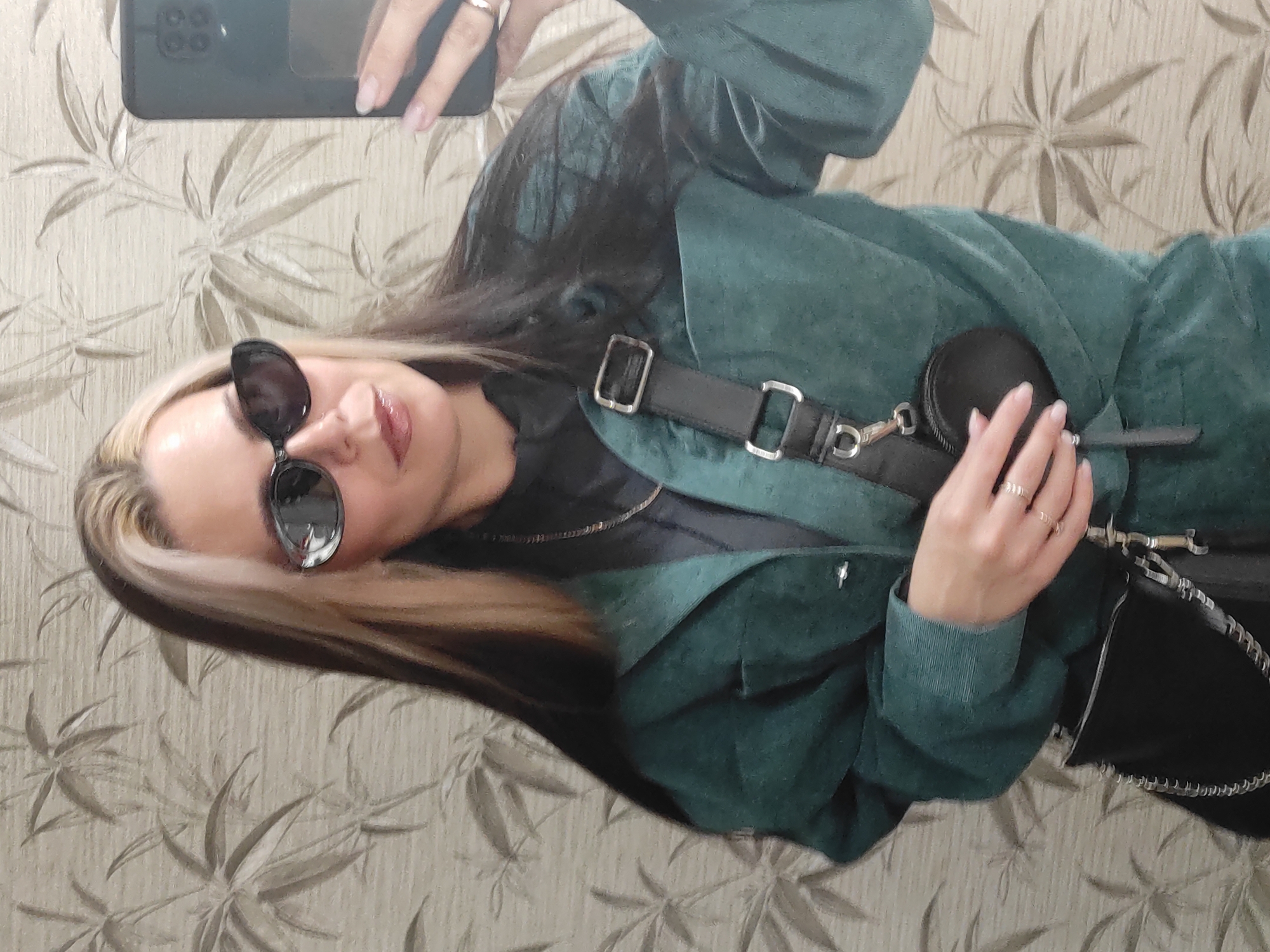 The image is a rotated selfie, capturing a young woman from the chest up as she holds a black smartphone to take the picture. She has long dark hair with distinctive blonde streaks framing her face and is wearing dark sunglasses that obscure her eyes. Her lips are slightly parted.
She is dressed in a dark teal or green jacket, which appears to be made of a textured material like corduroy or suede. Underneath, she wears a black top, and a delicate silver necklace is visible. A black cross-body bag with prominent silver buckles and hardware is slung over her shoulder, and a small, round black pouch is attached to its strap. Her left hand, adorned with several silver rings on her index, middle, and ring fingers, is visible resting near her bag. Her fingernails have a natural-looking, light-colored polish.
The background is a wall covered in wallpaper featuring a subtle, repeating pattern of light-colored, stylized leaf or floral shapes on a neutral beige or cream background. The scene takes place indoors, likely within a private residence in Chaikovsky, Russia, under ambient indoor lighting. No visible text is clearly discernible.