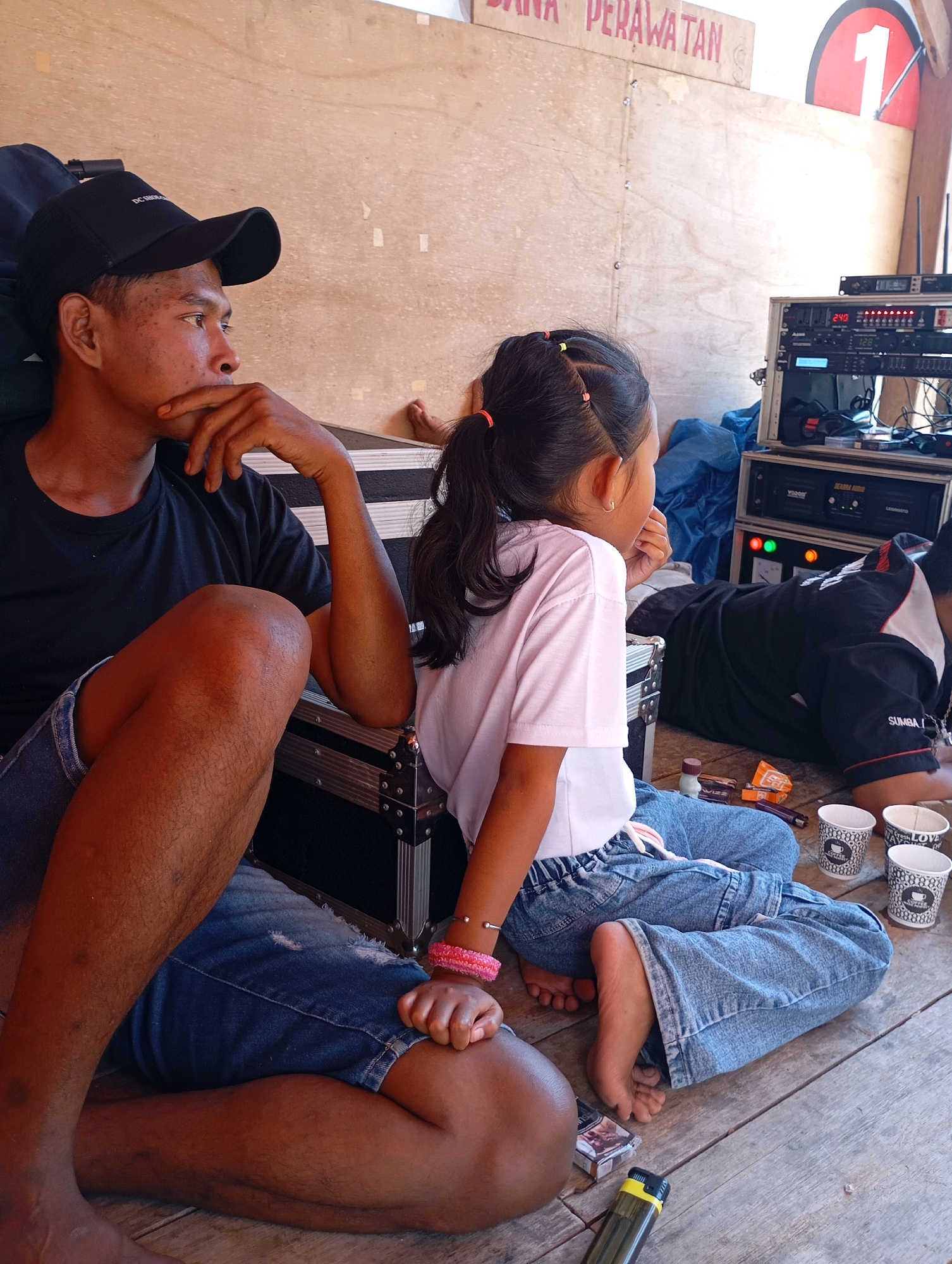 In Ketepung, Indonesia, a man wearing a black t-shirt and a black baseball cap sits on a wooden floor with his legs crossed, his hand resting on his chin as he looks to the side. Beside him, a young girl in a pink t-shirt and blue jeans also sits on the floor, her back to the camera. In the background, several pieces of audio equipment are stacked on top of each other, and a large wooden panel with text is visible on the wall. The scene appears to be outdoors, possibly at an event or performance space, given the presence of the audio equipment.