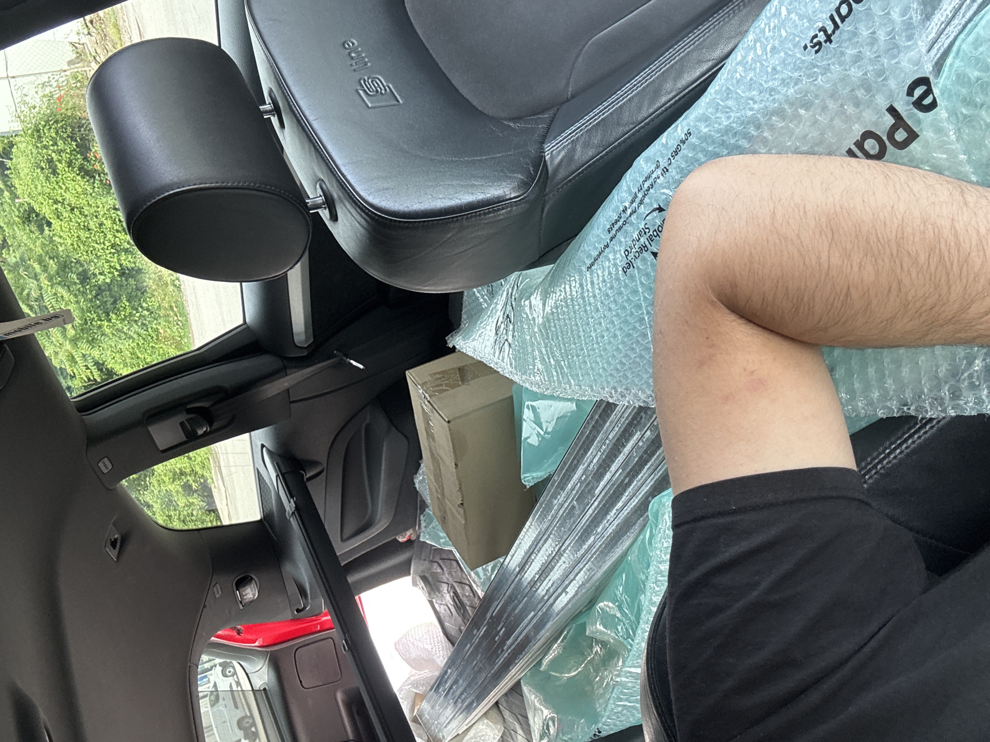 The image depicts the interior of a car, likely a hatchback, filled with various items. A person's leg, wearing a black t-shirt, is visible in the foreground, resting on a large roll of blue bubble wrap. The car's black leather seats, featuring the "S line" logo, are prominent. In the back, several packages and long, metallic objects are visible, some wrapped in protective materials. The car appears to be parked outdoors, with greenery and a fence visible through the windows, suggesting a suburban or semi-rural environment. The context provided indicates the location is the city of Shumen, Bulgaria.