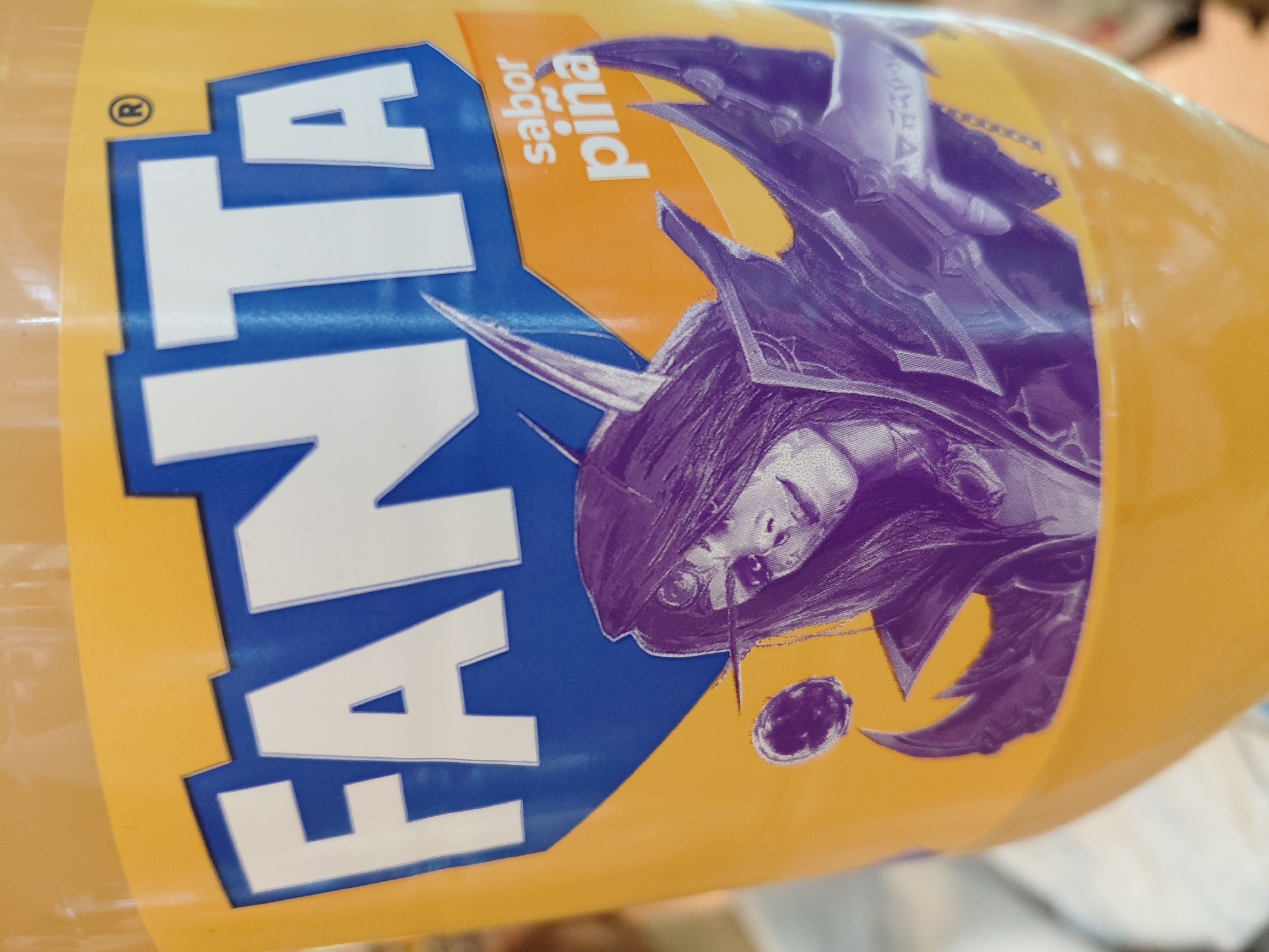 This is a close-up, eye-level shot of a Fanta pineapple-flavored soda bottle, showing the iconic Fanta logo and a character illustration. The Fanta logo, in white letters outlined in blue, is prominently displayed across the top of the bottle. Below the logo is a detailed, purple-toned illustration of a female character with elven ears, ornate armor, and what appears to be a mystical orb in the upper left portion of the frame. The character's facial features are distinctly rendered with fine lines. To the right of the illustration, a yellow label with the words "sabor piña" (pineapple flavor) is visible. The bottle itself is made of translucent yellow plastic, through which the beverage inside can be seen. The lighting is bright, suggesting an indoor setting, possibly a store or home. No specific location in Encarnacion, Paraguay, is identifiable from the image.