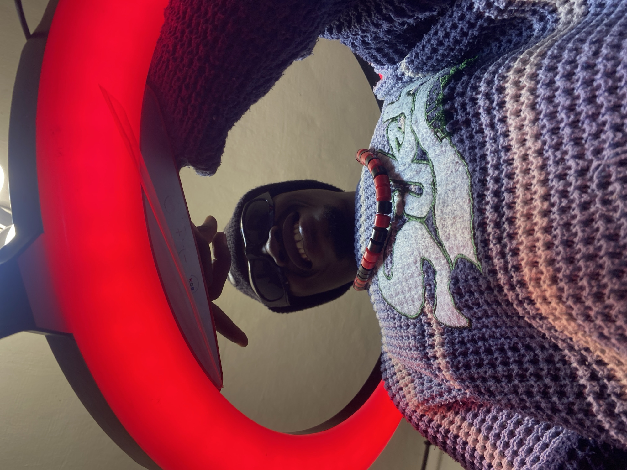 The image is a low-angle, close-up shot of a person, likely male, wearing a textured purple and pink knitted garment with a light blue embroidered design. The person is also wearing sunglasses and a dark beanie. A necklace with red and black beads is visible around the neck. The person is holding a device with a red illuminated ring light, possibly a camera or phone accessory, and appears to be pointing at the device with their right index finger. The background is a plain, neutral-colored wall. The image is taken from a perspective looking upwards at the subject, suggesting the camera is positioned low on the ground or held from below. There are no clear indicators of the specific location within Abakaliki, Nigeria, but the indoor setting and modern lighting equipment suggest an urban environment.