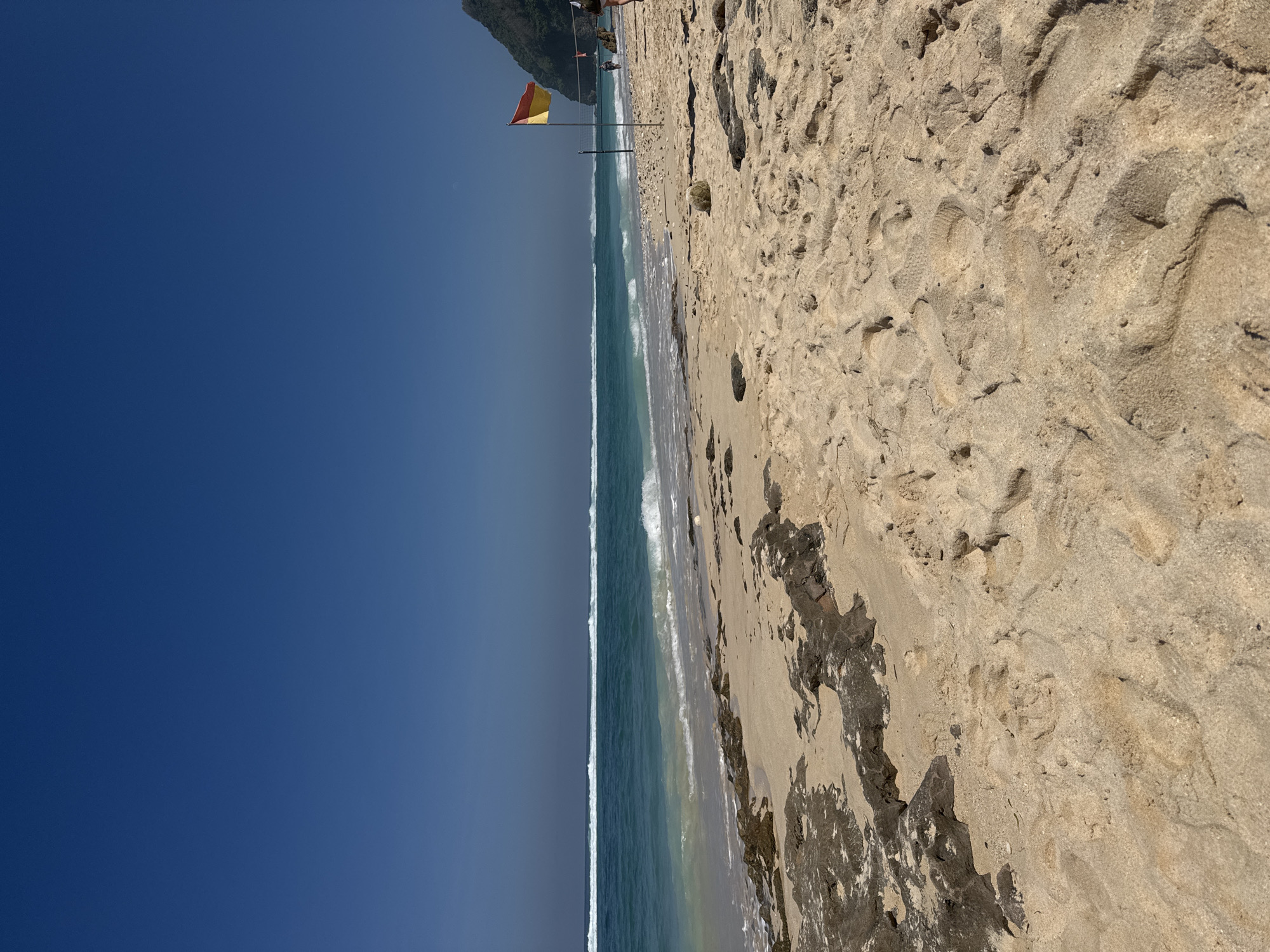 The image depicts a bright, clear day at a sandy beach with a vast, deep blue sky dominating the upper portion. The sea, with a distinct turquoise hue near the shore transitioning to deeper blue further out, features multiple parallel lines of white breaking waves extending horizontally across the mid-ground. The sandy beach in the foreground and mid-ground is light beige, textured with numerous footprints and irregular indentations. Dark, irregular rock formations are visible along the shoreline on the left and scattered within the sand in the immediate foreground.
On the right side of the beach, a yellow and red rectangular flag is attached to a vertical pole. Adjacent to and slightly left of the flag, a volleyball net structure with poles and a net is discernible. A light-colored, spherical object, possibly a deflated ball, rests on the sand in the mid-foreground, to the left of the flag. Further to the right, near the base of a steep, dark green, vegetated cliff or hill that rises dramatically, two human figures are present on the sand; one appears to be walking, and another is partially visible at the extreme right edge of the frame. The scene is brightly illuminated, indicating direct sunlight.