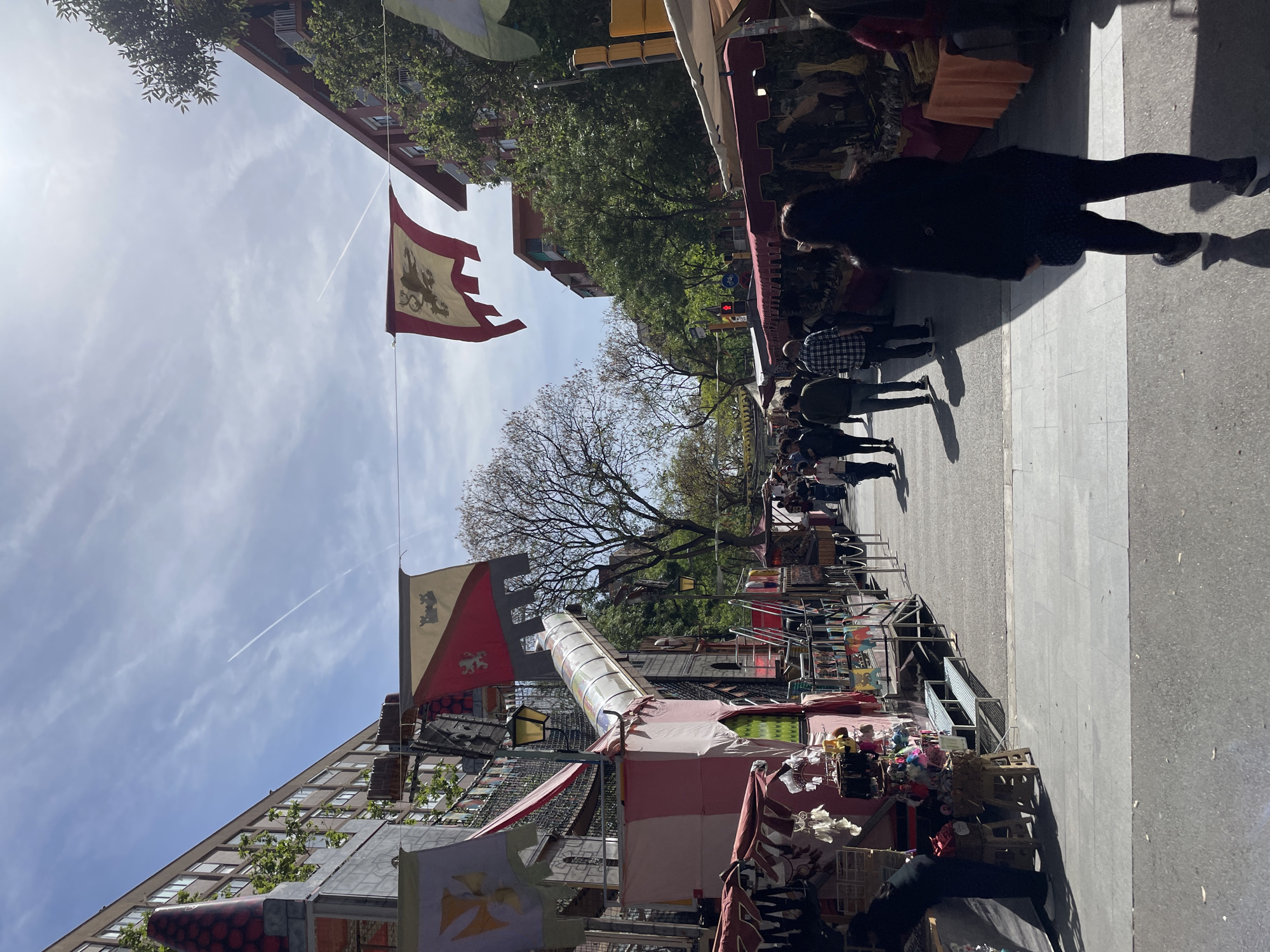 The image captures an outdoor market or fair taking place on a sunny day in Barcelona, Spain. The scene shows a bustling street lined with numerous market stalls, with people strolling along, browsing the various goods on display.
The market stalls feature colorful awnings and banners, many adorned with heraldic designs, including lions, castles, and crosses, suggesting a medieval or historical theme. Among the items visible at the stalls are textiles, crafts, and plush toys. On the right side of the street, a paved area serves as a walkway for pedestrians, while the street itself appears to be part of the market area.
The time of day is clearly daylight, with bright sunshine casting distinct shadows from people and structures, indicating mid-morning to early afternoon. The sky is bright blue with scattered clouds, and a white contrail from an airplane is visible across the expanse. Tall, leafy trees line parts of the street, and multi-story buildings can be seen in the background. A traffic light and streetlights are also present. The overall atmosphere appears lively and festive, with attendees engaged in the market activity. No specific text is clearly legible on any signs or banners.