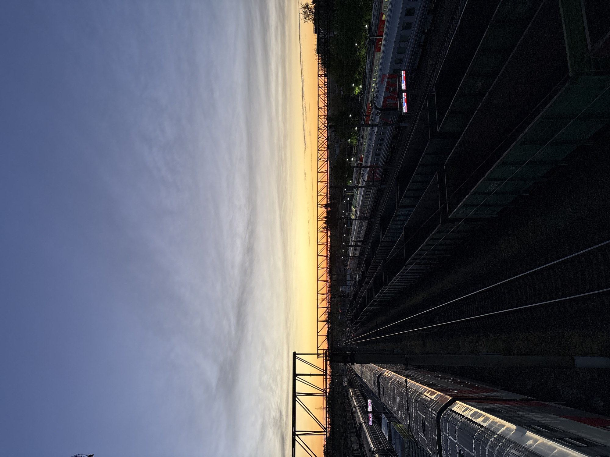 The image displays a railway scene oriented vertically, showing the sky at the top, a horizon line, and railway infrastructure extending below it.
**Subjects:**
The upper portion of the image is dominated by the sky, which transitions from a deep blue at the very top to a soft, luminous yellow and orange hue along the horizontal horizon line, indicating twilight. Wispy, light clouds are scattered across the sky. A prominent, dark metallic overhead gantry or bridge structure stretches horizontally across the middle of the frame, silhouetted against the bright horizon. Below this, numerous parallel railway tracks recede into the distance. Several passenger trains with multiple carriages are visible on the tracks, particularly on the right side. One train clearly displays an illuminated sign. In the deep shadows on the far right, darker, box-like freight wagons are also present. Another train carriage is partially visible in the bottom-left corner, featuring reflective surfaces. Distant artificial lights, possibly from platform or station lighting, are discernible.
**Activity or event taking place:**
The scene depicts a static moment at a railway yard or station during twilight. No active human presence or specific event is directly observable; the focus is on the stationary trains and infrastructure.
**Notable details:**
The time of day is clearly either sunrise or sunset, given the striking gradient of warm colors near the horizon contrasted with the darkening blue sky. The presence of artificial lights indicates that it is dusk or dawn. The overall lighting creates a dramatic contrast between the illuminated horizon and the deeply shadowed foreground elements like the tracks and trains. The setting is unequivocally a railway environment, likely a major station or depot, consistent with the provided location context of Kaluzhskaya, Russia.
**Visible text:**
A prominent red and white logo, "РЖД" (RZD) for Russian Railways, is visible on the side of one of the passenger trains. Faint, dark numbers and letters, such as "332 467" and "0305574", are barely discernible on the shadowed freight wagons.