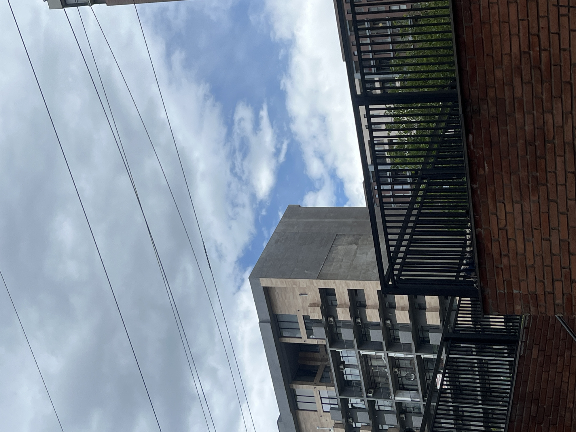 The image shows a view in Tbilisi, Georgia, looking upwards towards buildings and the sky. Electrical wires are strung across the left side of the frame, contrasting with the cloudy blue sky. A modern building with a mix of gray and tan facades occupies the central area, displaying several windows and possible HVAC units on its exterior. A brick building with a black metal balcony or fire escape runs vertically along the right side of the image. Vegetation is seen on the balcony, adding a touch of green.