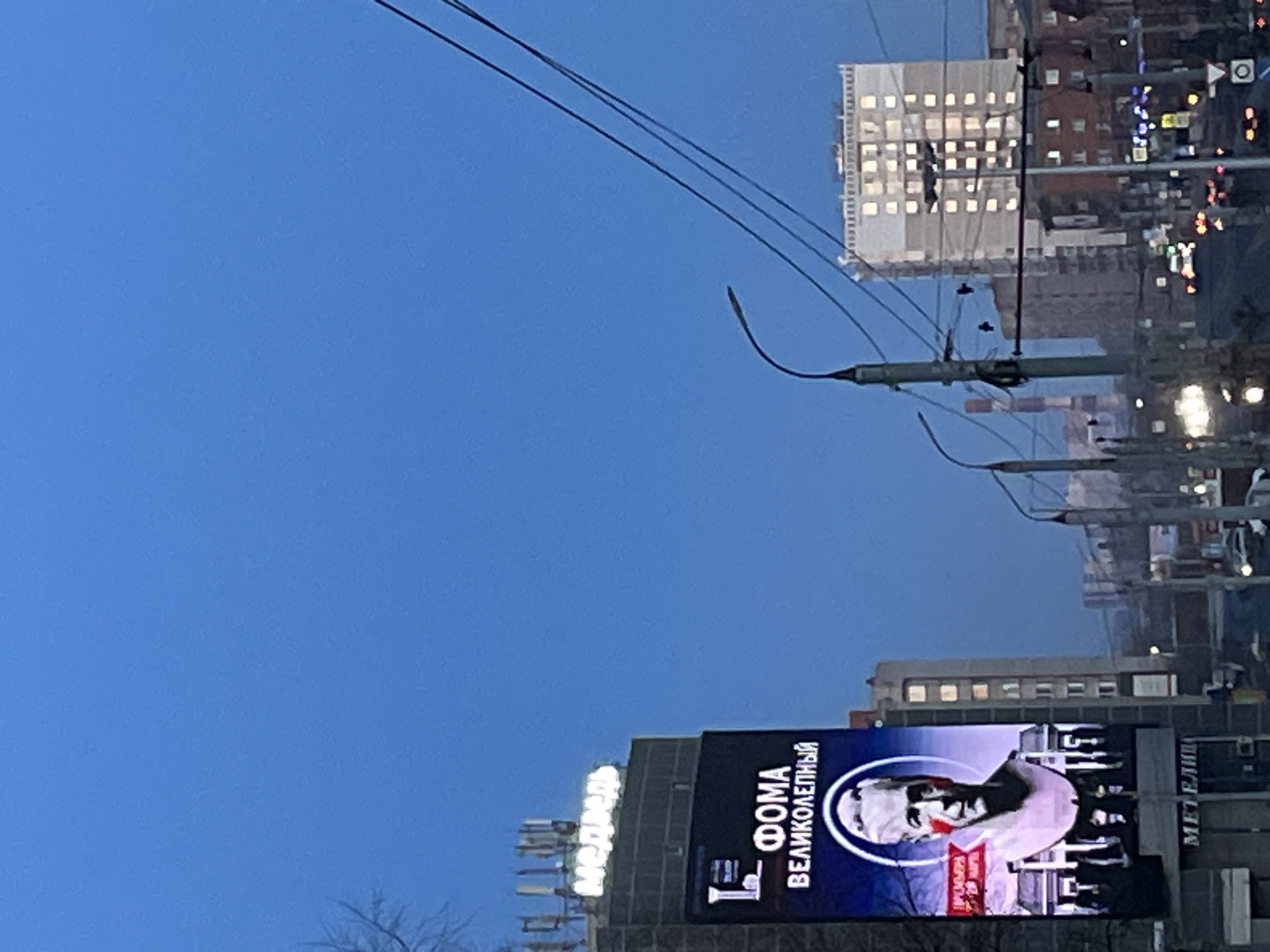 The image captures an urban scene, possibly in the city of Izhevsk, Russia, given the Cyrillic text on a large electronic billboard. The billboard displays an image of a person, presumably a significant figure, accompanied by the words "ФОМА ВЕЛИКОЛЕПНЫЙ" which translates to "THOMAS THE MAGNIFICENT".
The environment is a cityscape at dusk or early evening. Several buildings are visible, some with illuminated windows, suggesting business or residential activity. A network of utility poles and wires is strung across the sky. The sky itself is a deep blue.
The scene appears to be captured from street level, possibly from a vehicle or pedestrian viewpoint. The overall atmosphere is urban and slightly blurred due to the low light conditions.