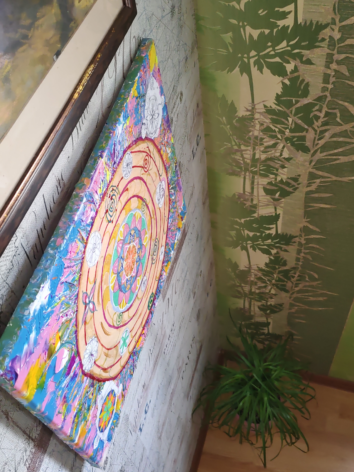 This is a photograph taken at an angle, focusing on a piece of abstract art with a mandala design, hung on a textured wall. The artwork is vibrant, featuring a central circular pattern with concentric rings and intricate floral motifs in various colors like orange, pink, blue, and yellow. The sides of the canvas are thickly painted with a palette knife, creating a textured, impasto effect in a riot of colors.
The wall behind the artwork has a subtle pattern, appearing to be wallpaper with a faint script or map-like design. To the right of the artwork, a portion of a green-toned wallpaper with a repeating stencil pattern of stylized trees or plants is visible. In the lower right corner, a potted green plant with long, thin leaves sits on a light wooden floor, near the corner of the room. The lighting in the scene is moderate, highlighting the textures and colors of the artwork.
The photograph does not provide any visual cues that would allow for the identification of its location in the city of Rostov-on-Don, Russia.