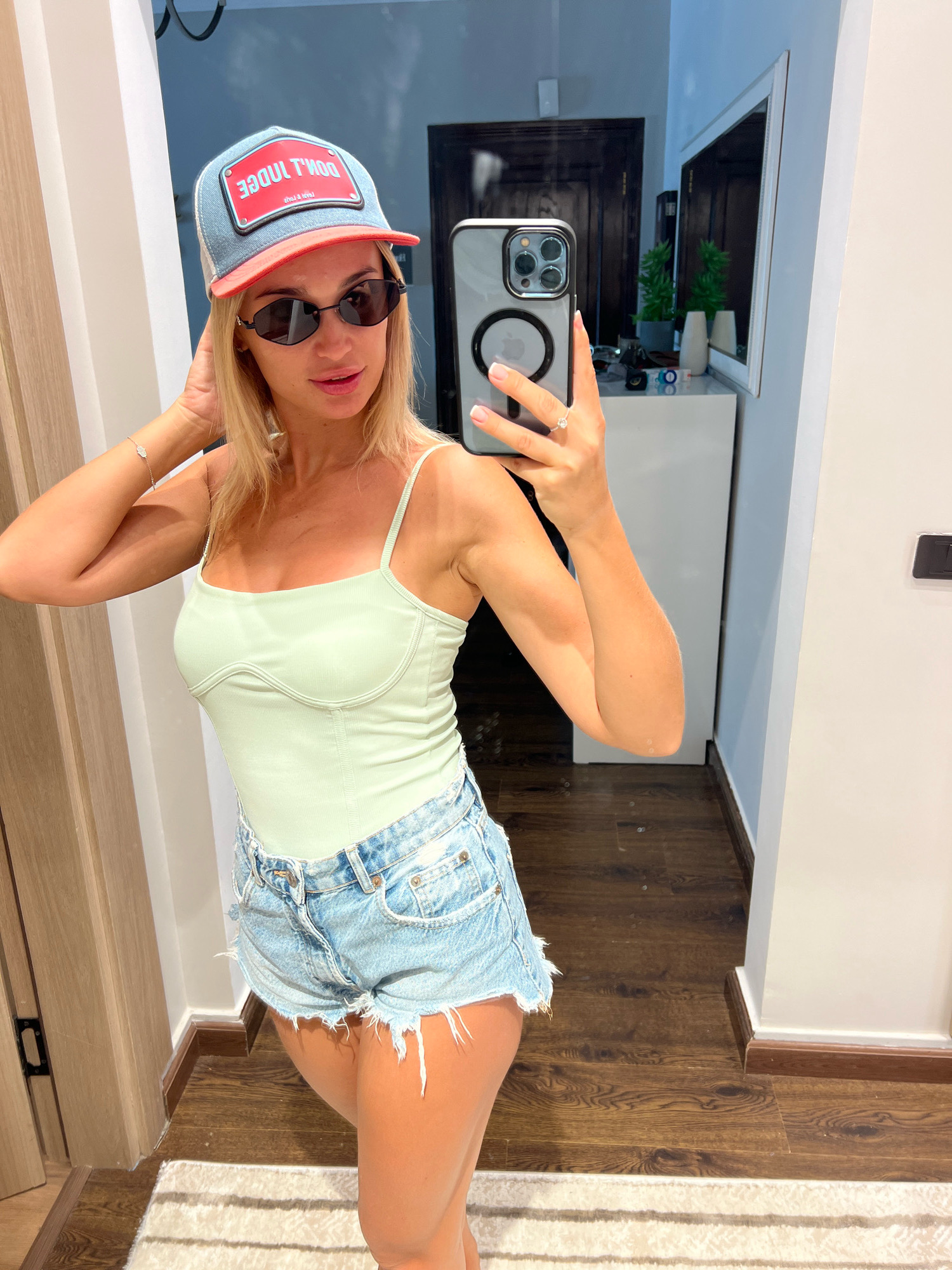 A blonde woman is taking a mirror selfie in what appears to be a bedroom or dressing area. She is wearing a light green camisole, distressed denim shorts, sunglasses, and a blue and red baseball cap. She is holding her phone up to take the picture, and her left hand is touching her hair. The room has wooden floors and a striped rug. In the background, there is a dresser with plants and toiletries. The overall impression is casual and relaxed, suggesting a personal moment captured in a private setting.