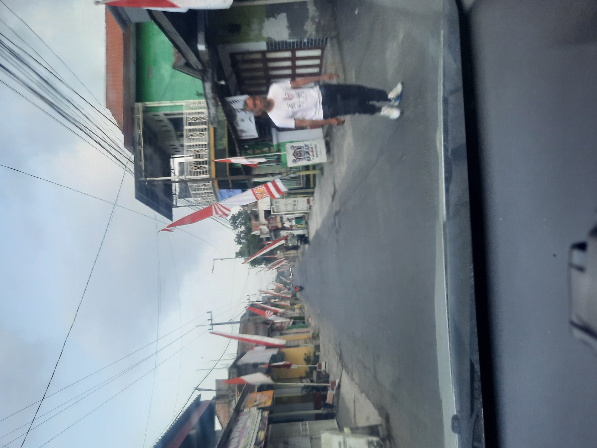 The image depicts a street scene in Banjarkemantren, Indonesia, lined with buildings adorned with numerous red and white Indonesian flags, suggesting a national holiday or celebration. A man wearing a white t-shirt and dark pants walks on the right side of the street. The street is paved and appears to be a local thoroughfare with some traffic, including a motorcycle visible in the distance. Overhead, a tangle of power lines crisscrosses the sky, which is overcast. The perspective of the image suggests it was taken from inside a vehicle, looking out through the windshield. Shops and houses are visible on both sides of the street.