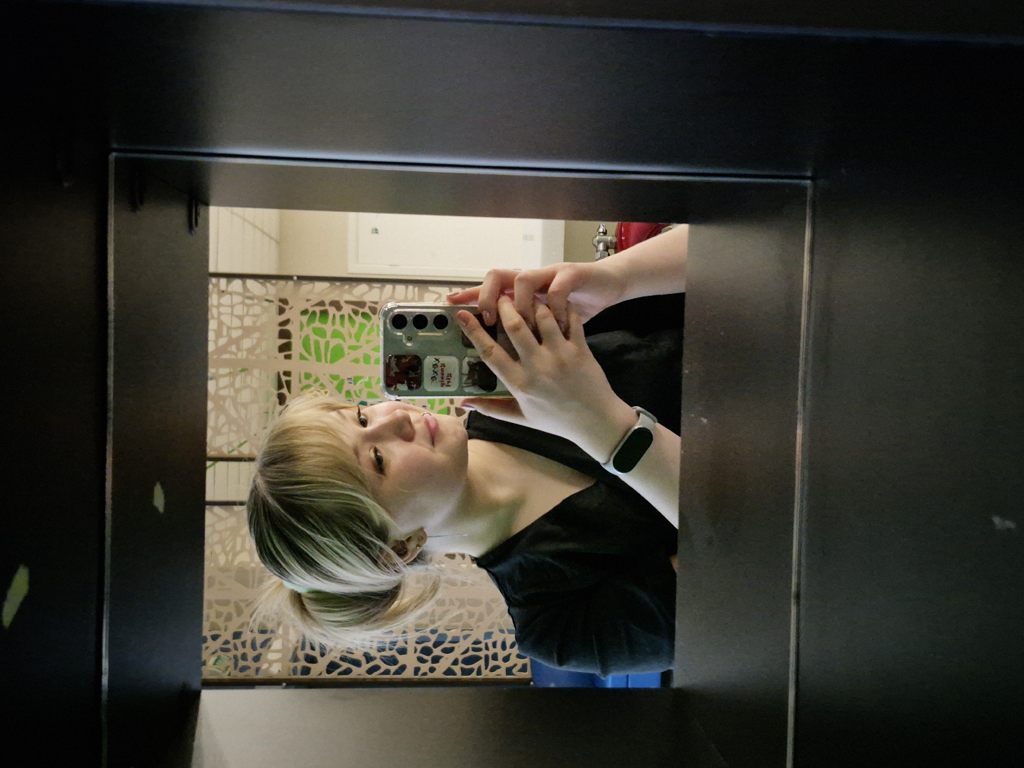A young person, likely female, takes a mirror selfie in an indoor setting within Chaikovsky, Russia. She has blonde hair with visible green or turquoise highlights/tips, pulled back into a low ponytail. Dressed in a black top, she wears a white smartwatch on her left wrist. She holds a smartphone with a clear case in both hands to frame the shot. The phone case features several stickers, including "xo xo," "I <3 Friends," and one depicting red lips.
The subject displays a subtle, pleasant smile, looking directly into the phone's camera lens. The mirror has a dark, thick frame that borders the reflection on three sides. The background, visible in the mirror, shows a light-colored wall or decorative screen with an intricate, organic, or geometric pattern. Behind this pattern, a bright green surface is reflected. To the upper right, a red object, possibly a faucet or part of plumbing, is partially visible, suggesting a public restroom or washroom in a modern establishment. The lighting is even, indicating an indoor environment, likely during the day.