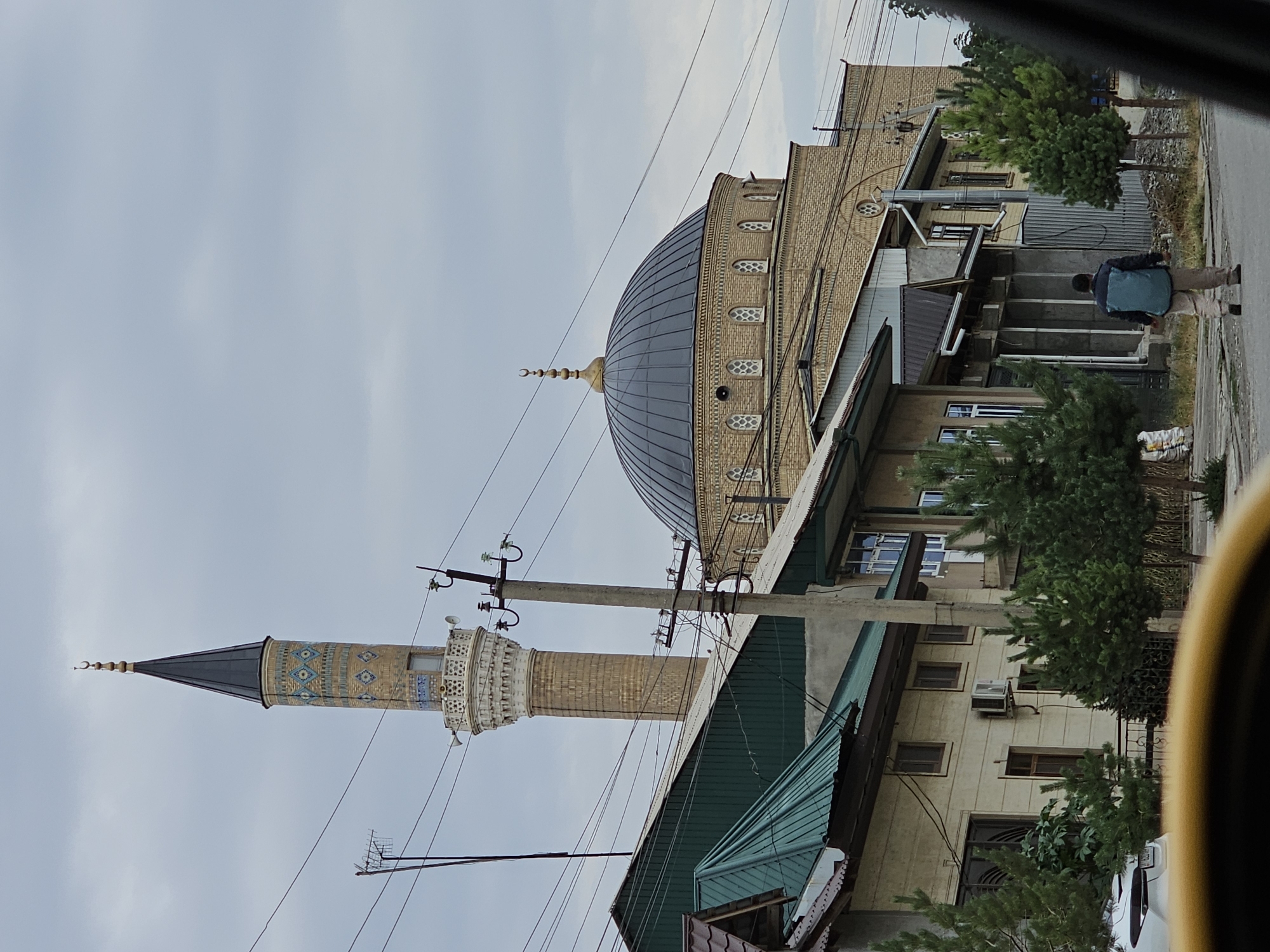 This is a street-level, medium shot photograph of a mosque in the city of Özgön, Kyrgyzstan. The mosque features a tall, slender minaret with intricate blue and beige tiling and a pointed black spire topped with a crescent moon. Adjacent to the minaret is the main body of the mosque, characterized by a large, dark-colored dome crowned with a smaller crescent. The mosque's facade is constructed of light-colored bricks, with arched windows and decorative architectural elements visible.
In the foreground, a row of residential buildings with various roof types, including green metal and darker corrugated materials, partially obscure the view of the mosque. These buildings have a more modest architectural style, with visible windows, air conditioning units, and exposed electrical wiring and utility poles. Evergreen trees are planted along the street in front of the buildings.
A single individual, a male wearing a blue and grey jacket and light-colored pants, is walking away from the camera down the street towards the mosque. A white vehicle is partially visible on the left side of the frame. The sky is overcast, suggesting a cloudy day. The overall scene depicts a blend of religious architecture and everyday urban life in Özgön.
