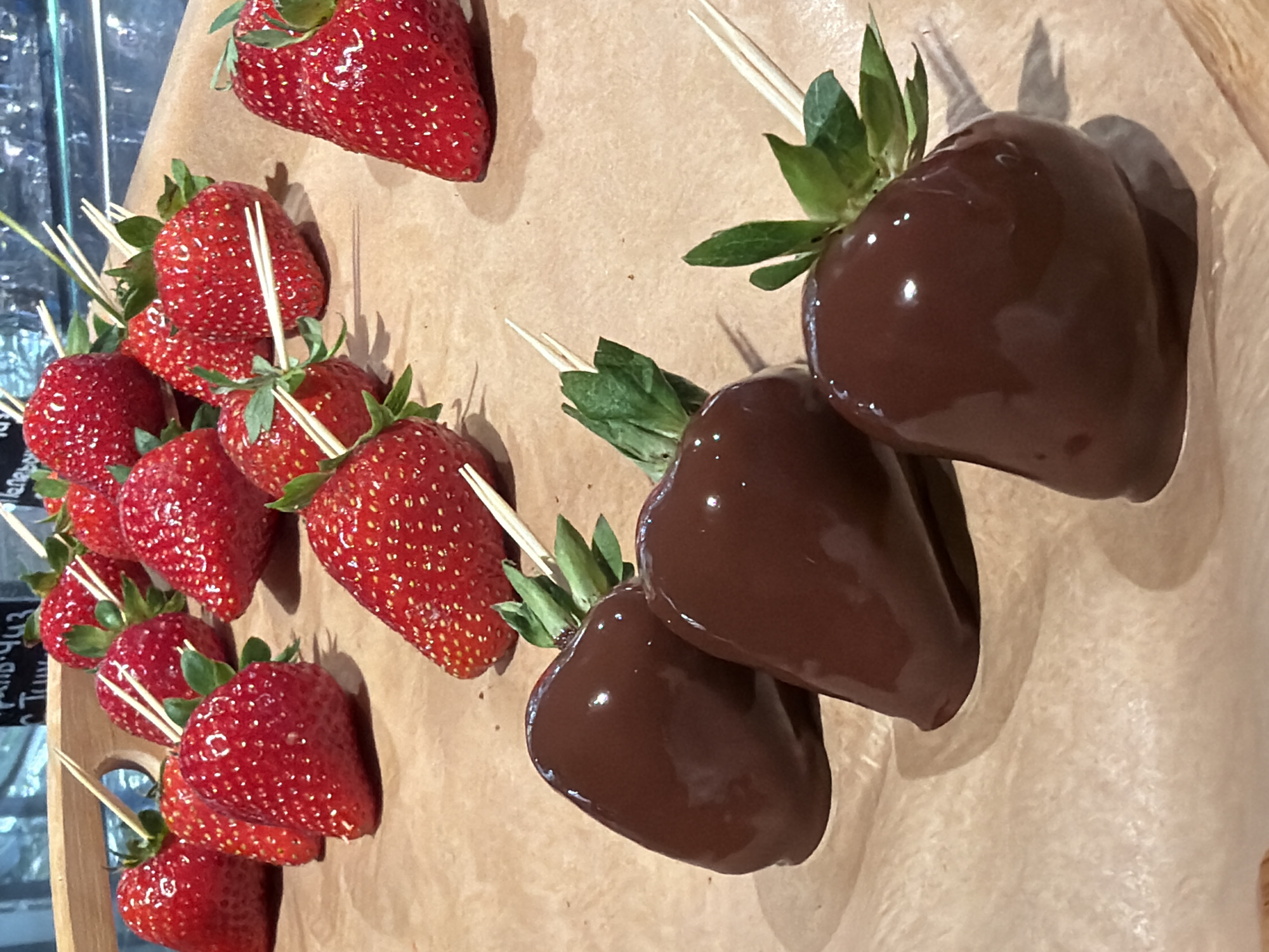 The image displays a close-up, eye-level view of several chocolate-dipped strawberries and plain strawberries arranged on a light brown surface, likely parchment paper, which is placed on a wooden surface. The strawberries are red and appear fresh. Some of the strawberries are whole and have been speared with small wooden sticks, while others have been dipped in dark chocolate, leaving only the green leafy tops exposed. The lighting suggests an indoor setting, possibly a bakery or confectionery shop in Saint Petersburg, Russia. The background is blurred, but hints of a counter and possibly display cases are visible. The composition focuses on the texture and sheen of the chocolate and the natural appearance of the fresh strawberries.