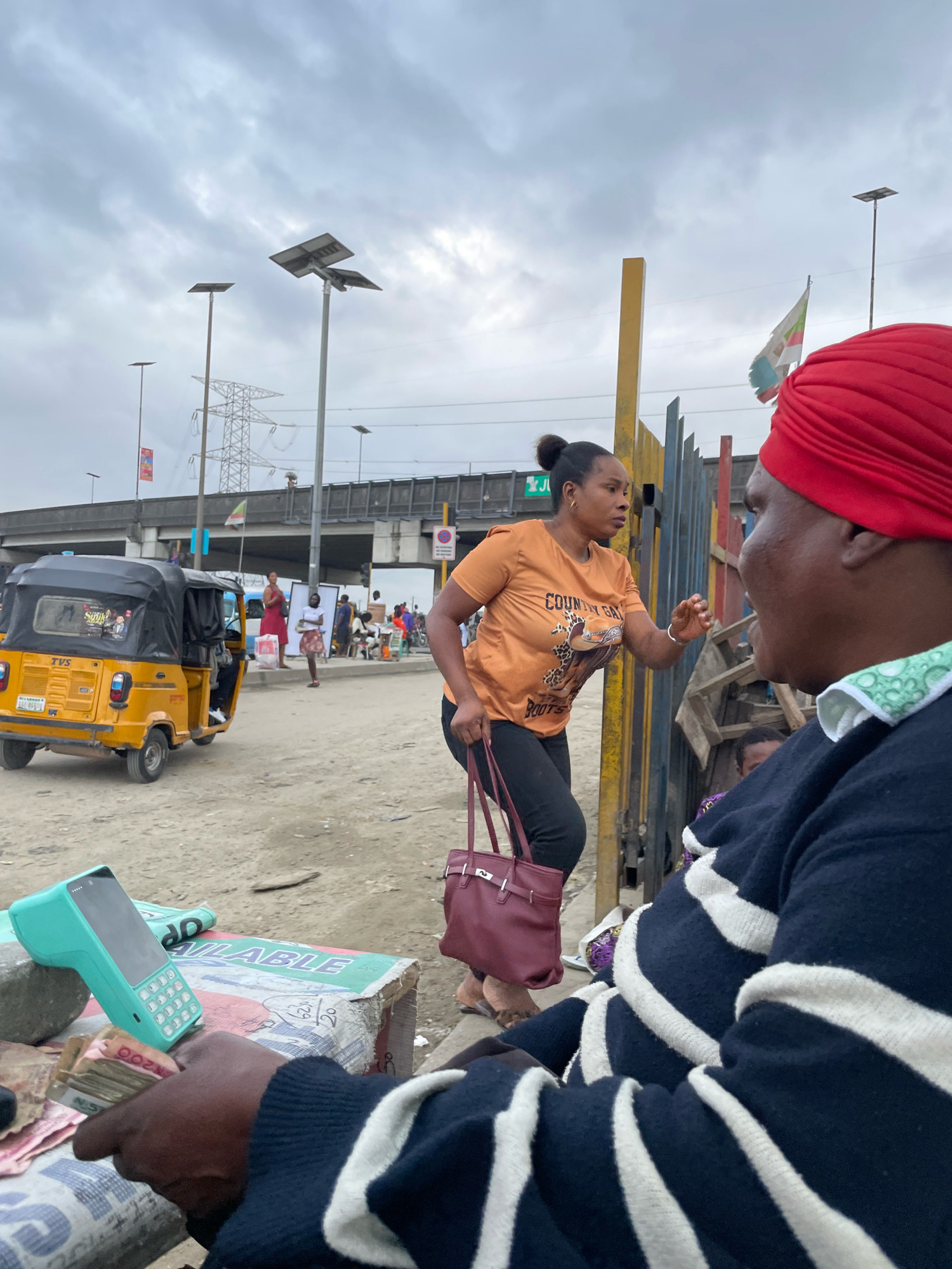 In Ajah, Nigeria, a woman in an orange t-shirt with "Country Girl Boots" printed on it, wearing black pants and holding a burgundy handbag, gestures emphatically towards another person whose face is obscured by a red headscarf and a navy and white striped sweater. This interaction takes place outdoors on a dusty street with a yellow auto-rickshaw parked nearby. In the foreground, a mint green card reader sits on a box, next to a stack of Nigerian Naira currency being held by a hand. In the background, several other people are visible, along with streetlights, power lines, and a metal barrier. The sky is overcast.