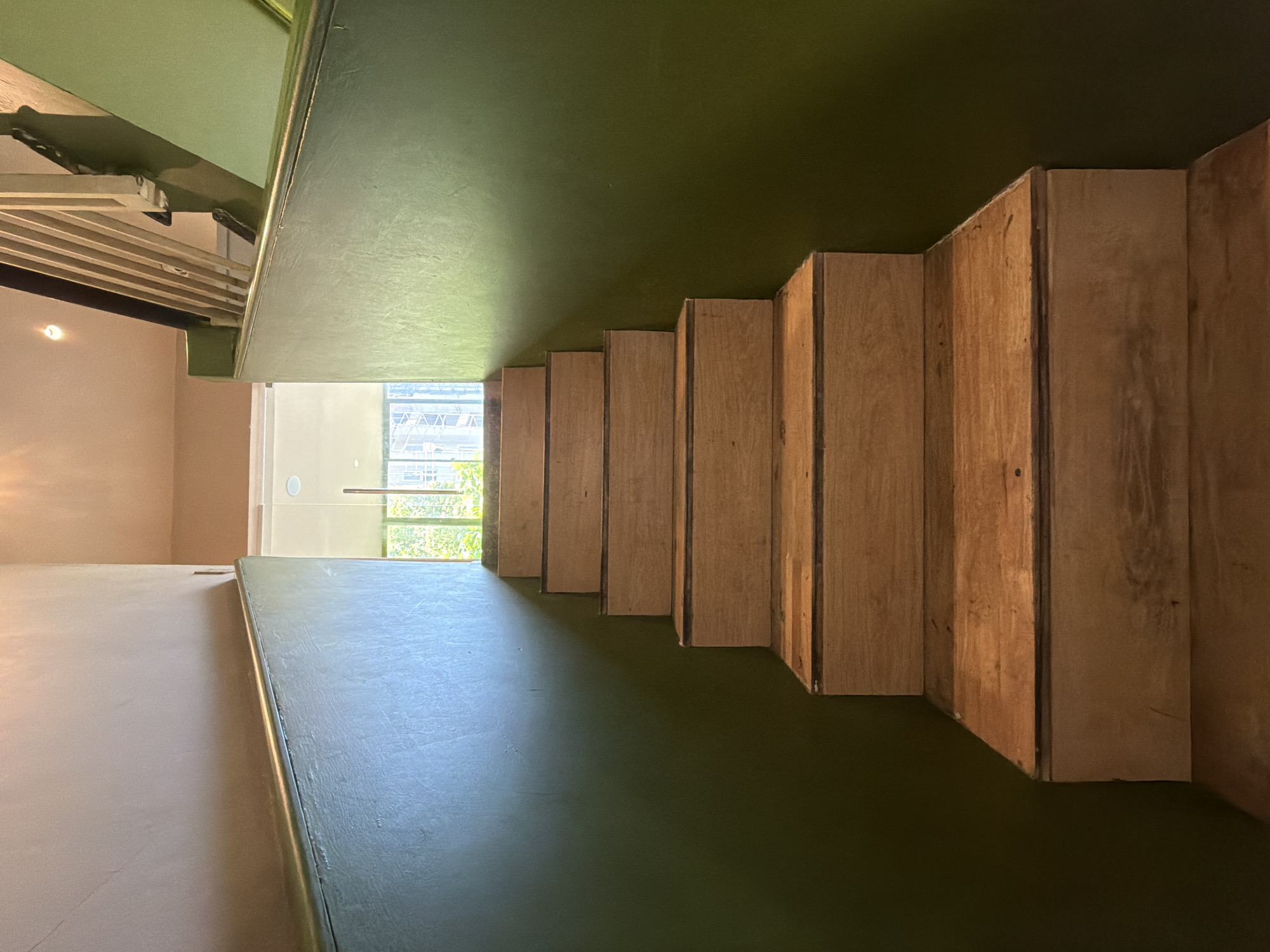 The image depicts a perspective view of a wooden staircase, leading upwards. The stairs are constructed from light-colored wood and have a simple, modern design. They are flanked by walls painted in a dark green hue. Natural light illuminates the scene from above and from an opening at the top of the stairs, where a glimpse of trees and buildings can be seen through glass. The overall impression is one of a clean and contemporary interior space, possibly a home or a commercial establishment. The scene is devoid of people or any specific actions taking place. The location in Nha Trang Ward, Vietnam, is not visually discernible from the image itself.