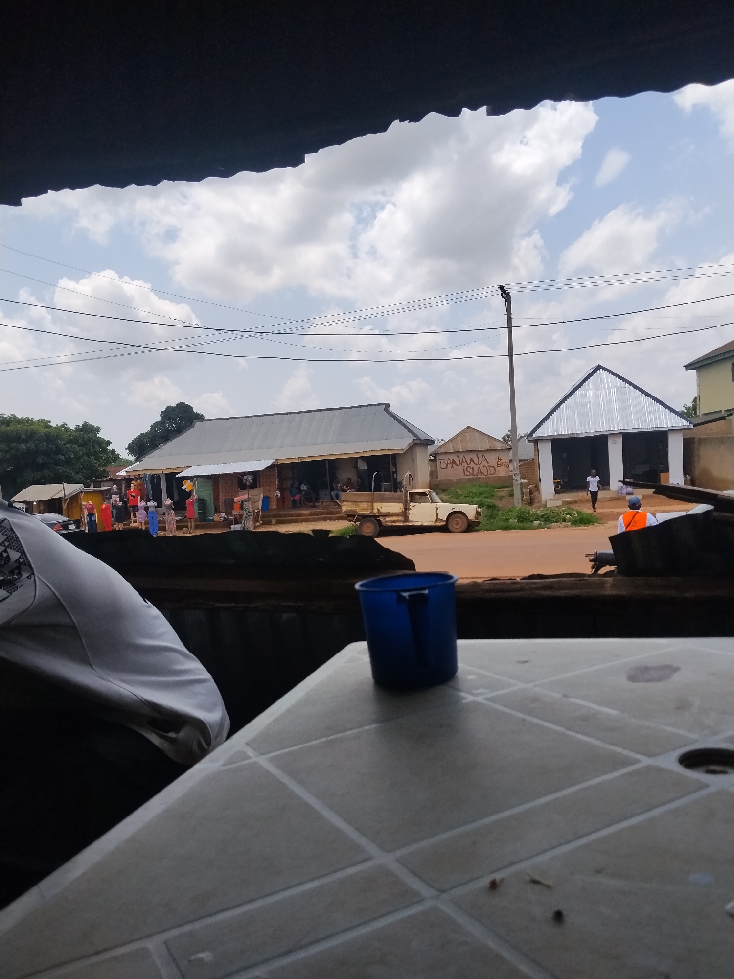 A street scene in Durumi, Nigeria, is captured under a cloudy sky. In the foreground, a blue plastic cup sits on a patterned table. In the middle ground, a dusty, light-colored pickup truck is parked on a red dirt road. To the right of the truck, a building with a corrugated metal roof and white walls is visible, with a person in an orange vest walking by. To the left of the truck, a row of shops with similar corrugated metal roofs lines the street. Mannequins dressed in various colorful clothing are displayed outside some of these shops. Trees and utility poles with electrical wires are scattered throughout the scene. The overall impression is of a bustling but somewhat unkempt commercial area.